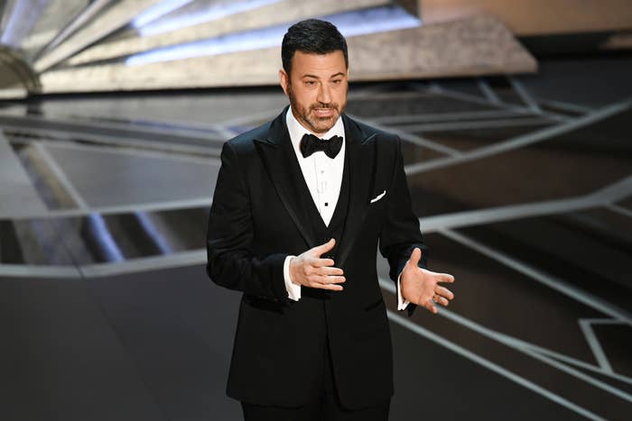 Jimmy Kimmel stands on stage in a black tuxedo, speaking with hand gestures