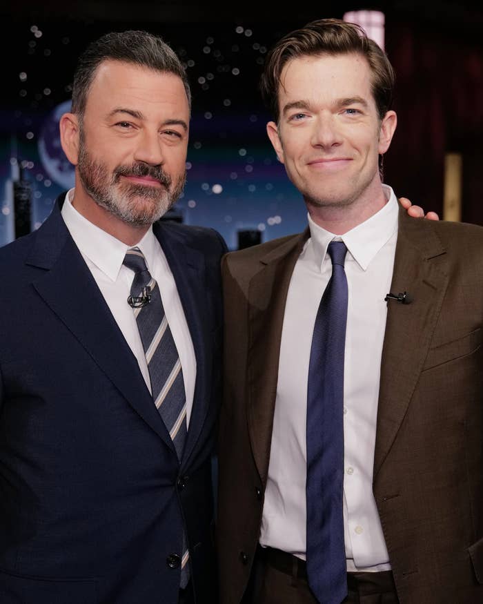 Jimmy Kimmel in a dark suit and John Mulaney in a brown suit pose together on a set
