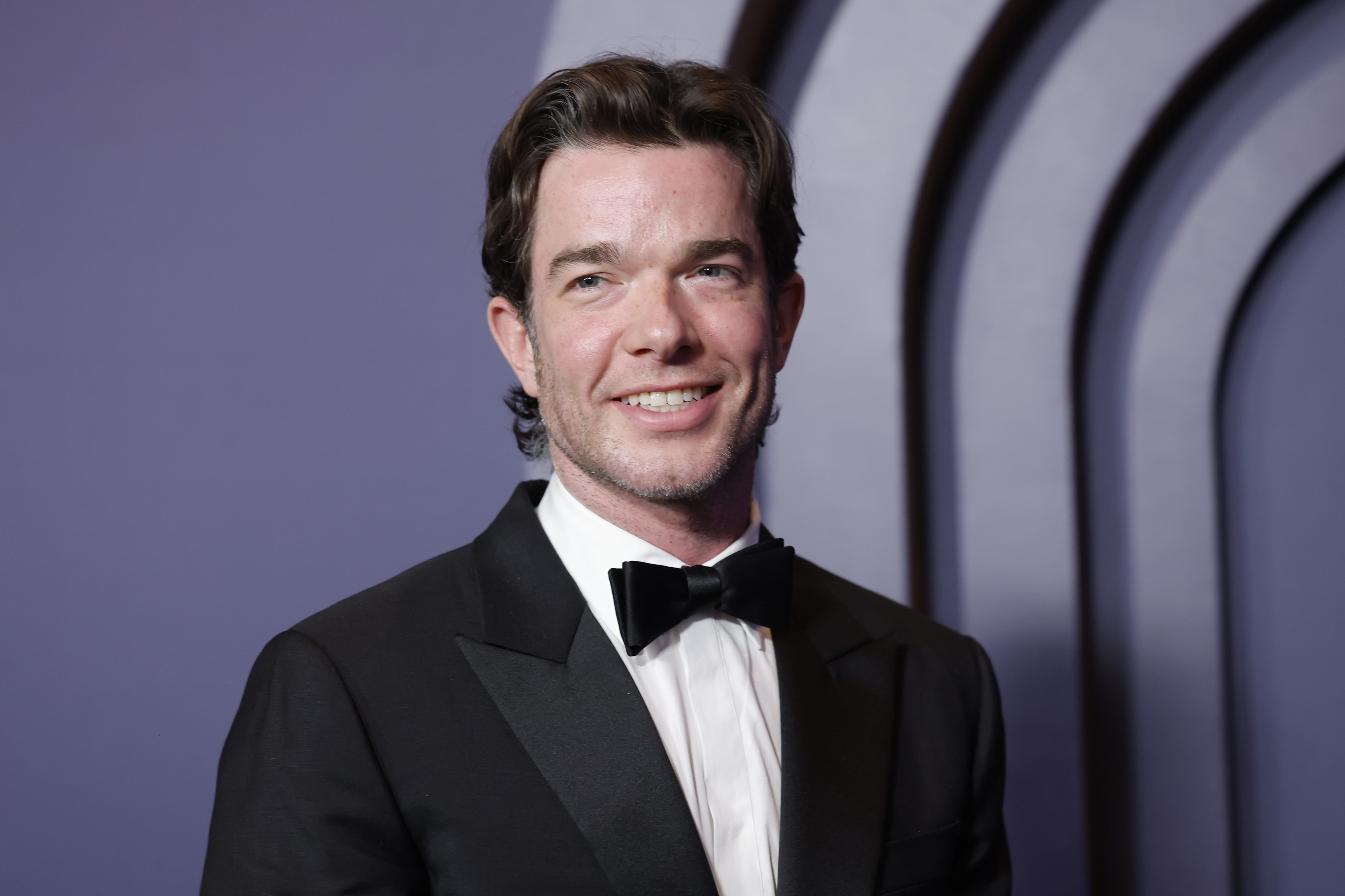 John Mulaney, in a black tuxedo with a bow tie, smiles while posing for a photo at an event