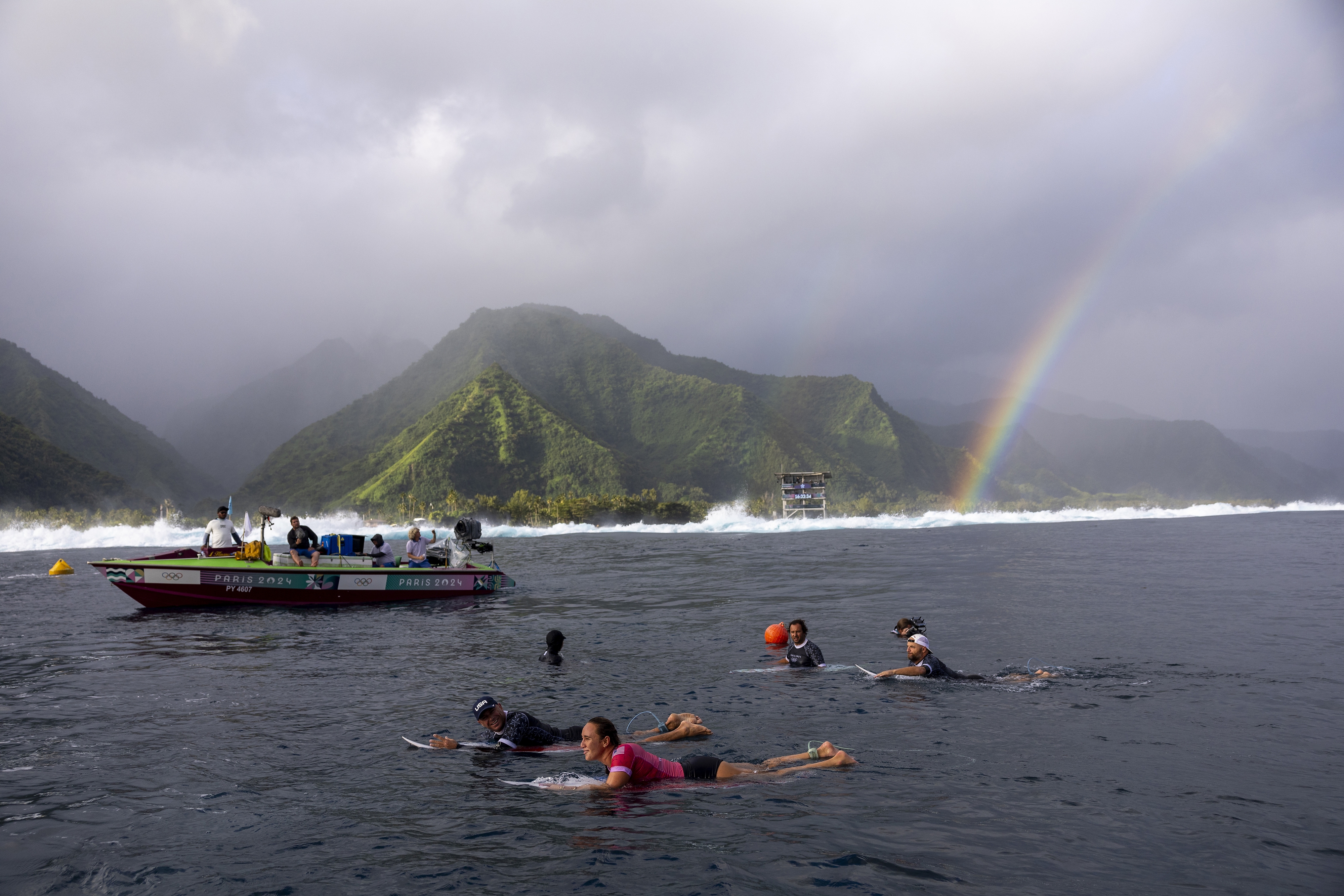 A group of people wearing wetsuits swim in the ocean near a boat with a rainbow and lush mountains in the background