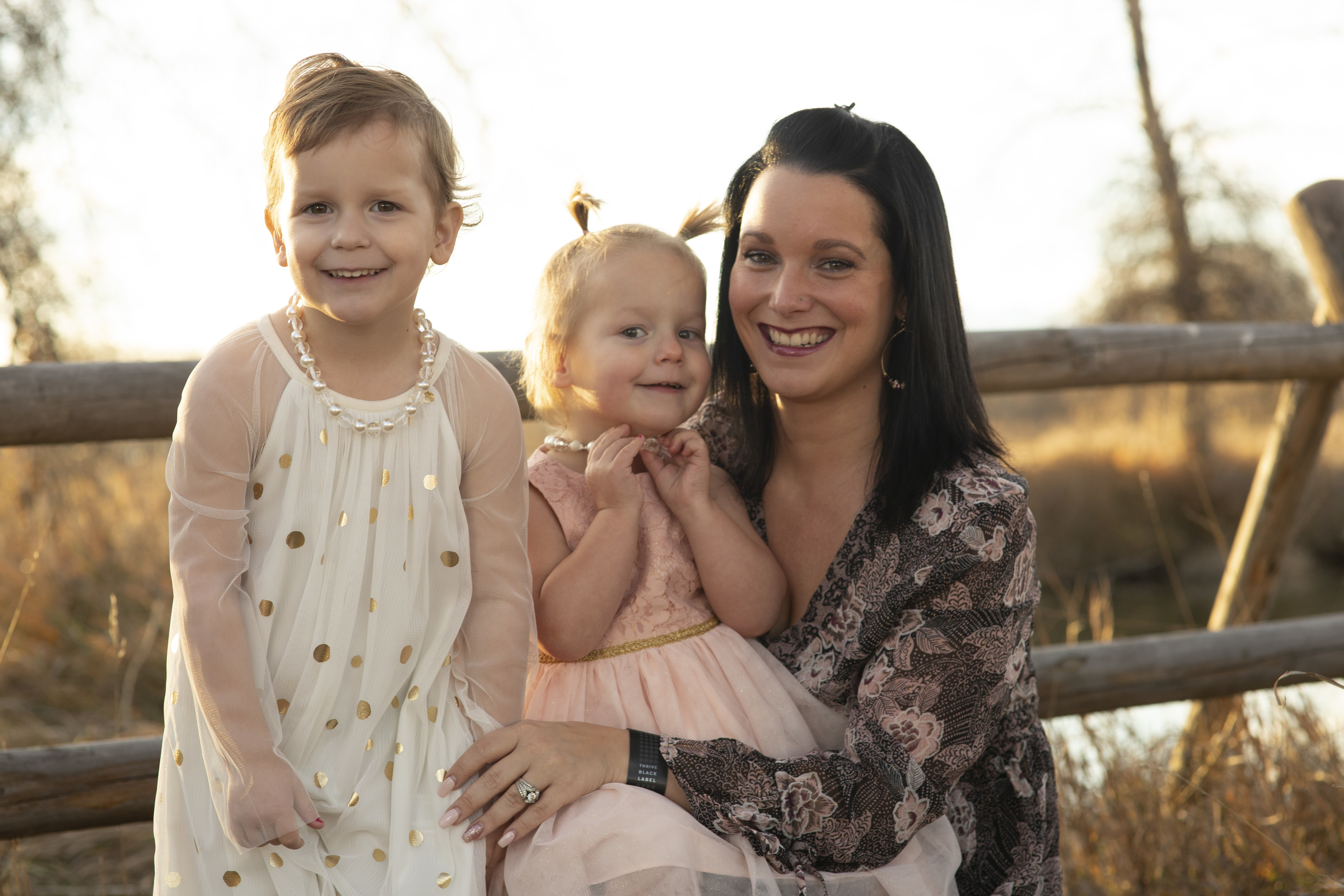 A woman, smiling and wearing a patterned dress, poses with two young girls, one in a netted gown with gold spots and the other in a simple dress. They're outdoors