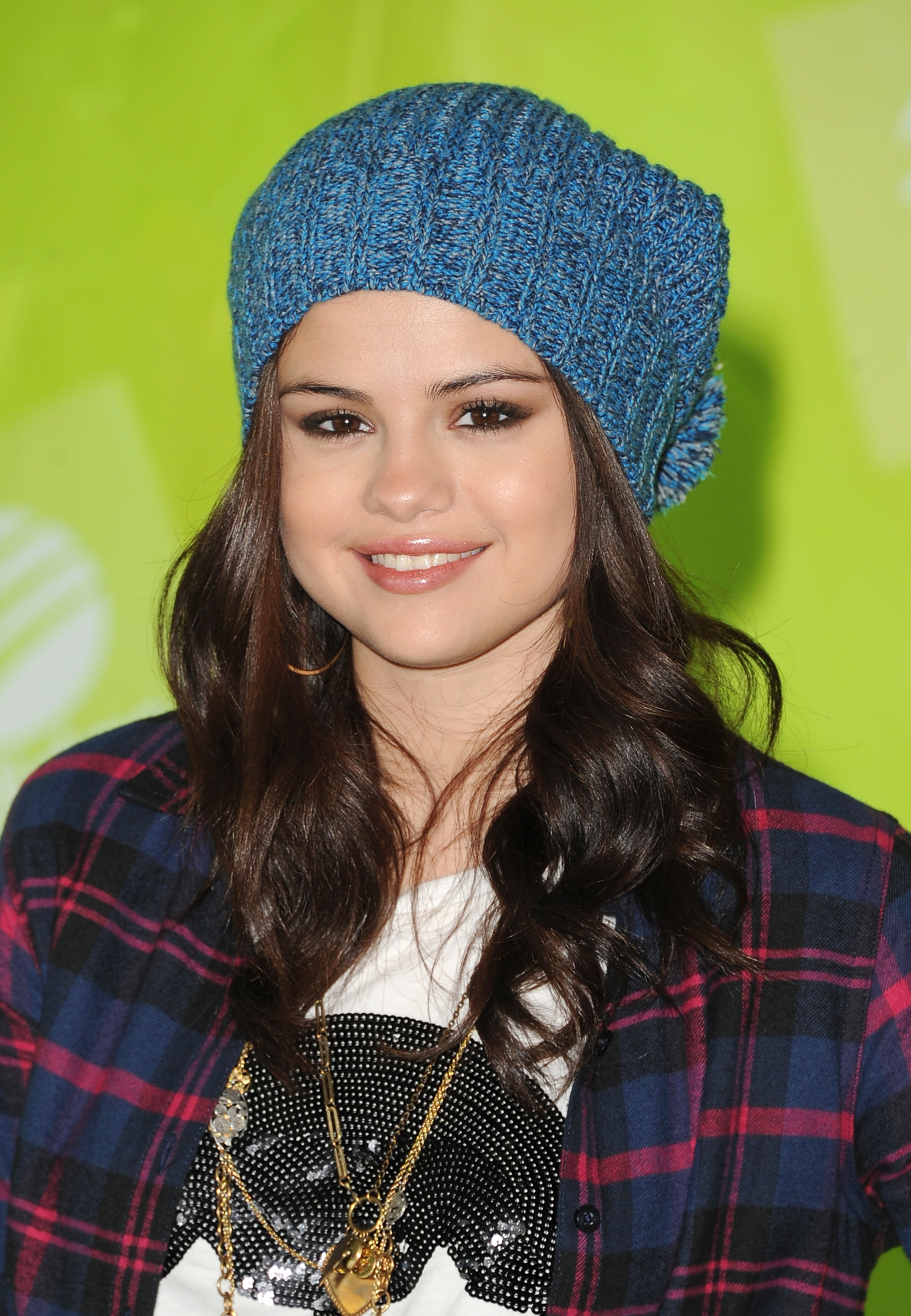 Selena Gomez smiling, wearing a knitted beanie, plaid shirt, and graphic tee, with layered necklaces
