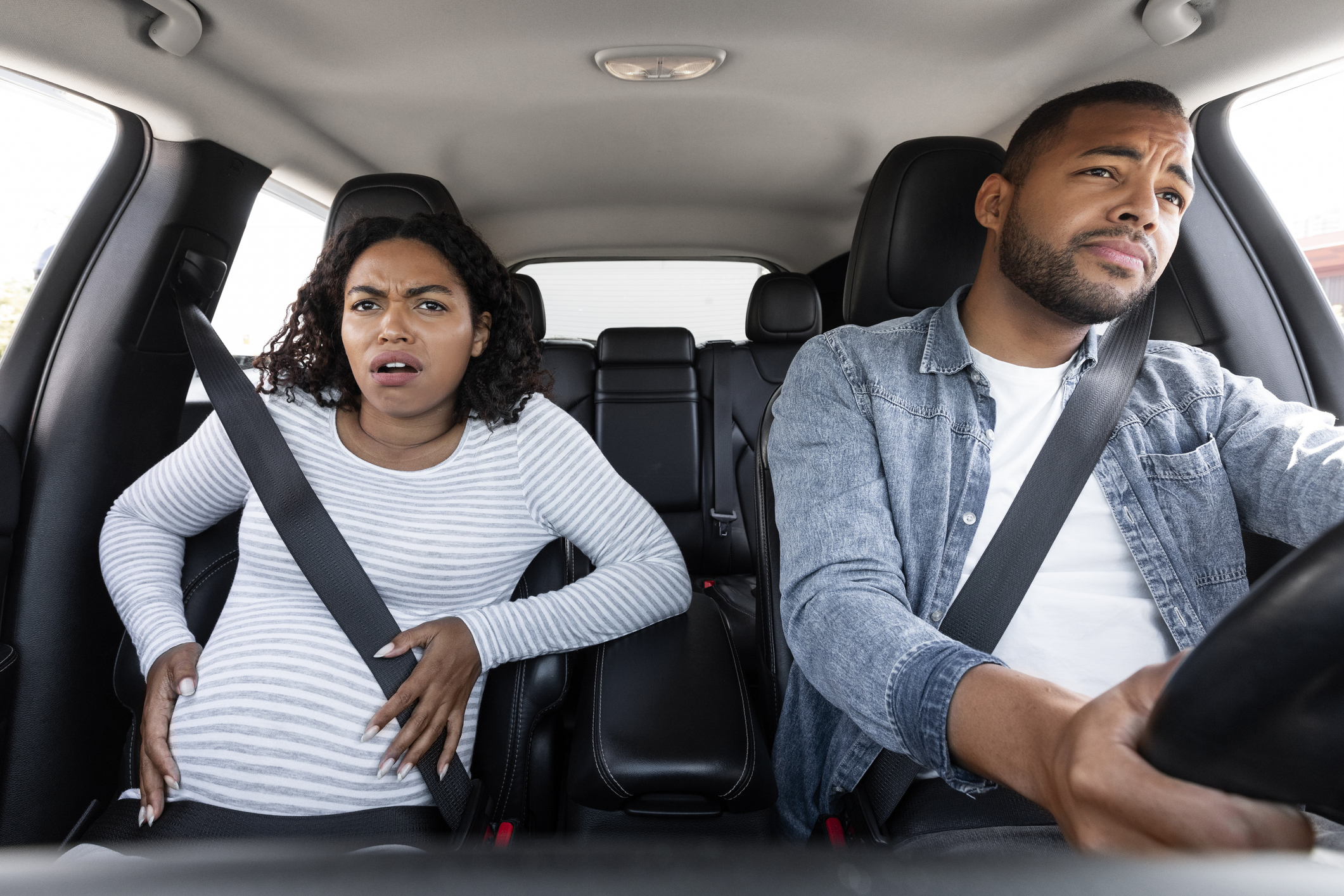 Pregnant woman in a striped shirt and man in denim jacket driving, both looking concerned