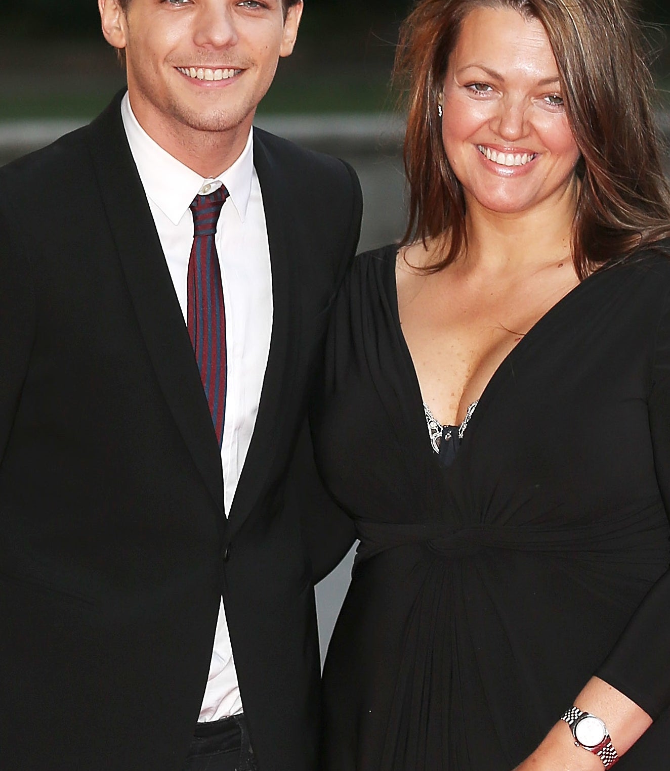 Louis Tomlinson in a black suit and tie, with his mother Johannah Deakin in a black dress and gold clutch, smiling together at a formal event