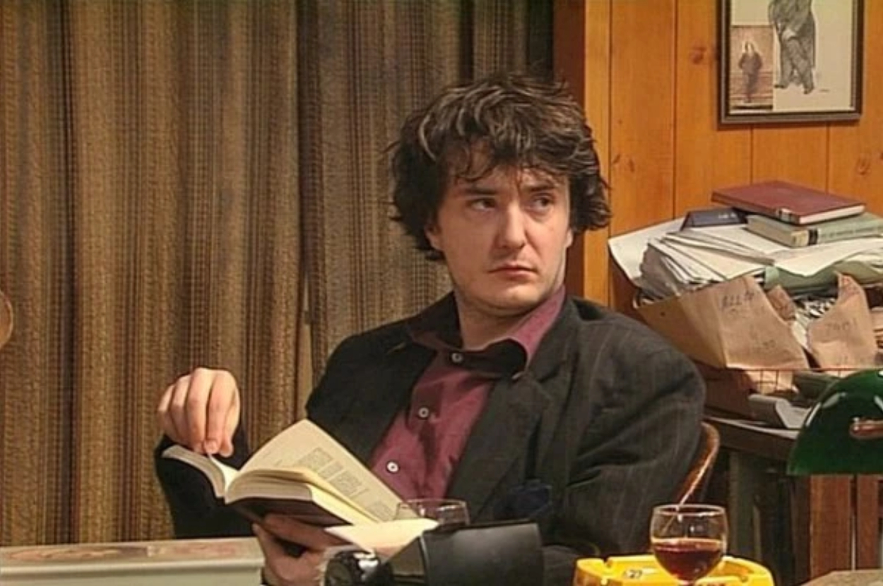Dylan Moran in character as Bernard Black from the TV show "Black Books," seated at a cluttered desk holding an open book and looking to the side