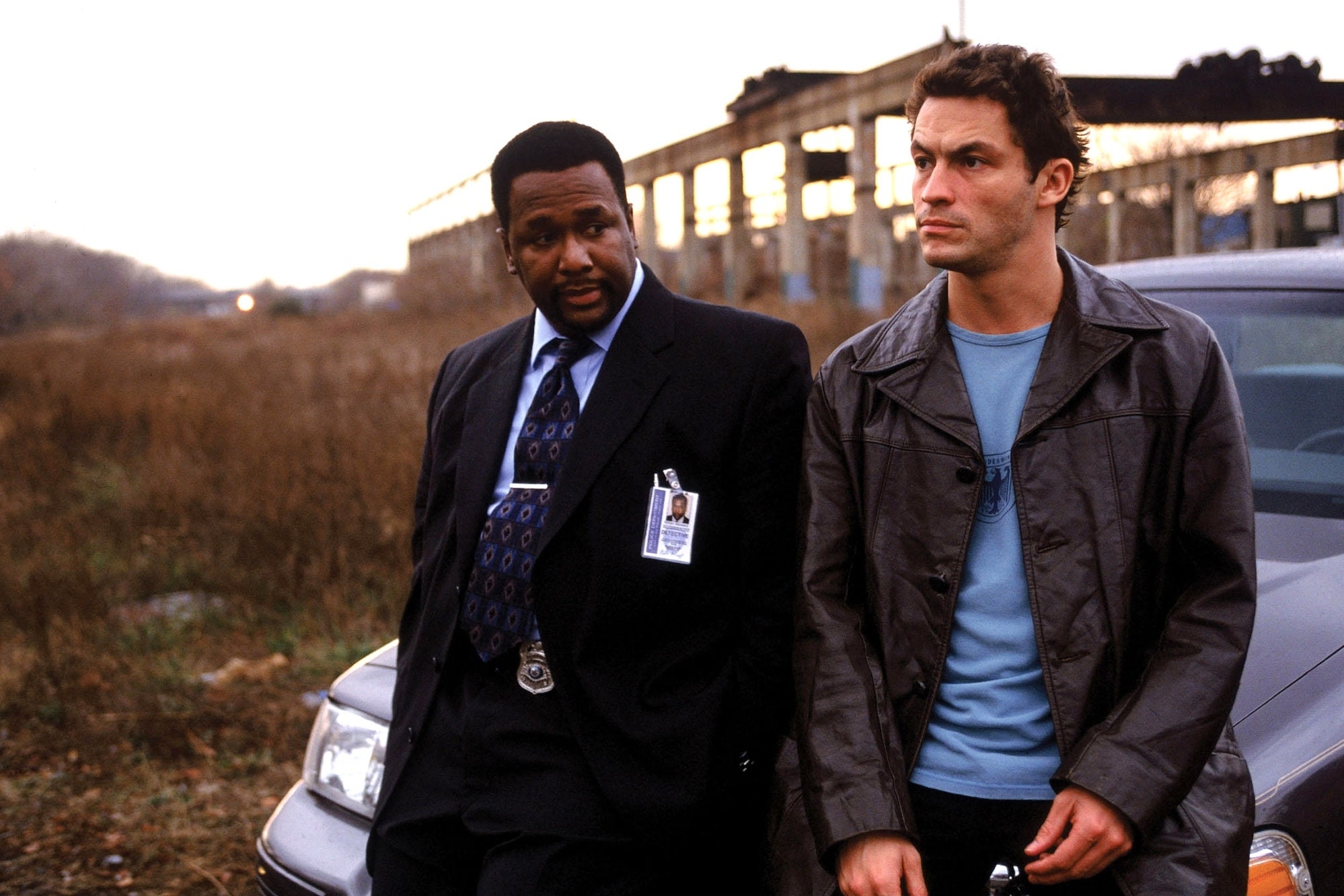 Wendell Pierce in a suit and badge, and Dominic West in a casual jacket, lean against a car with an industrial backdrop from "The Wire."