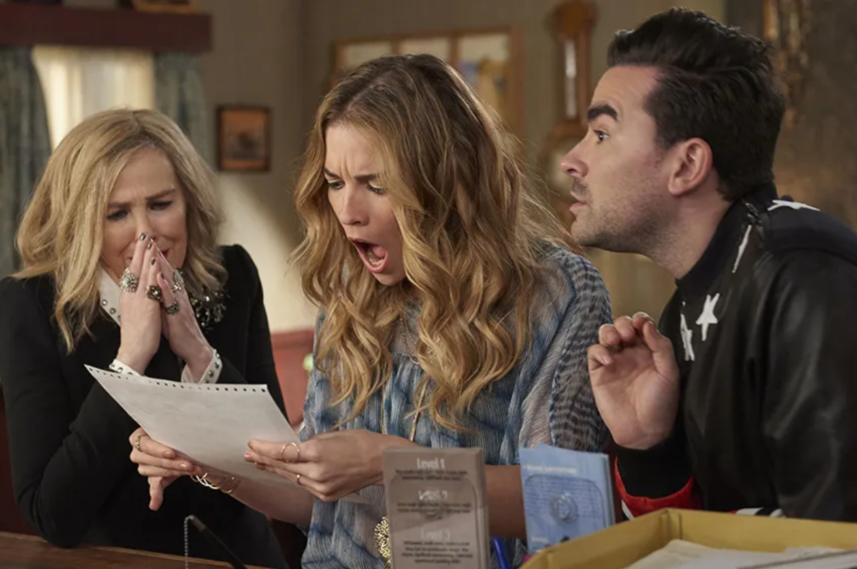 Catherine O'Hara, Annie Murphy, and Dan Levy are looking shocked at a piece of paper in a scene from a TV show