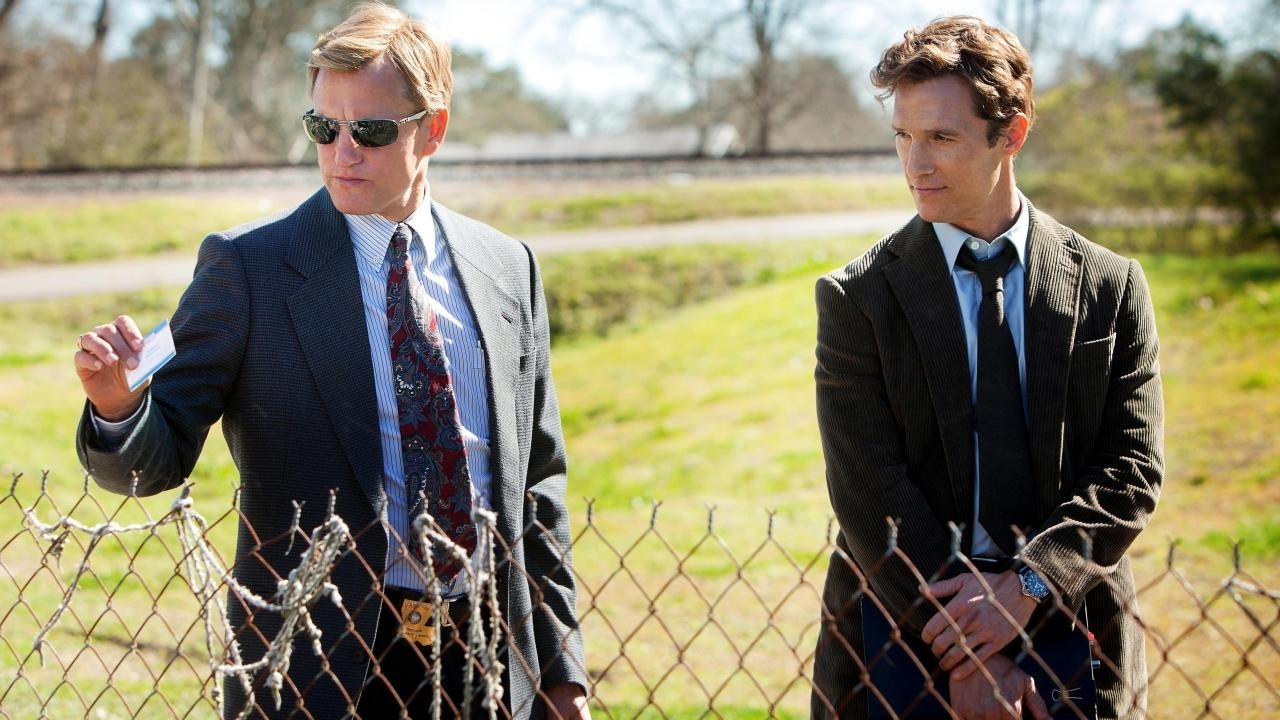 Woody Harrelson and Matthew McConaughey in suits, investigating a scene behind a fence, from the TV show "True Detective"