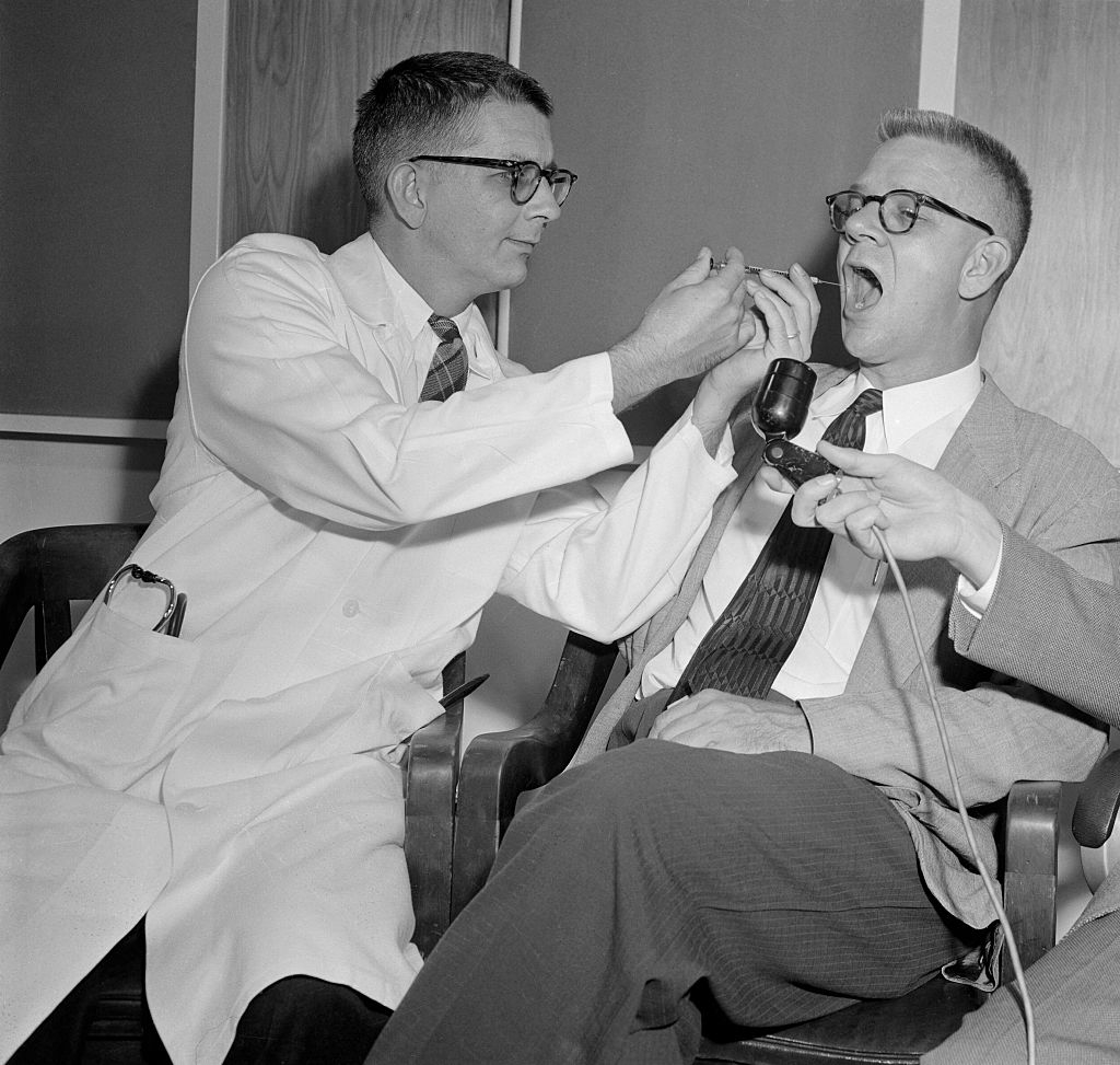 Two men in a medical exam, one in a lab coat checks the other's throat with a tongue depressor. They both wear glasses