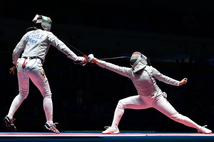 Two fencers compete; one labeled "Benitez VEN" lunges with sword while the other defends. Both wear full fencing gear, including masks and body protection