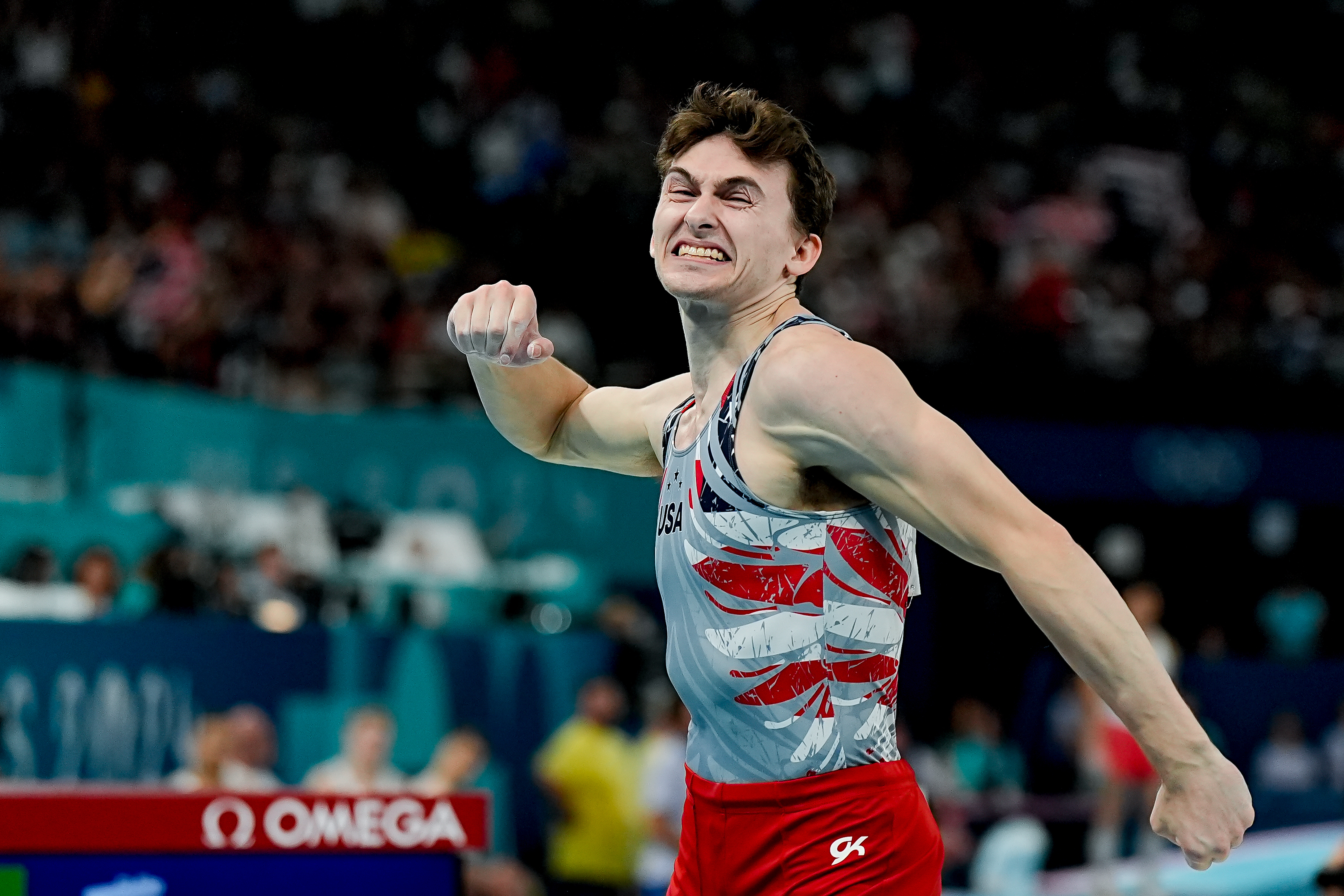 A gymnast celebrates a successful routine at a competition, flexing his arm with a triumphant expression
