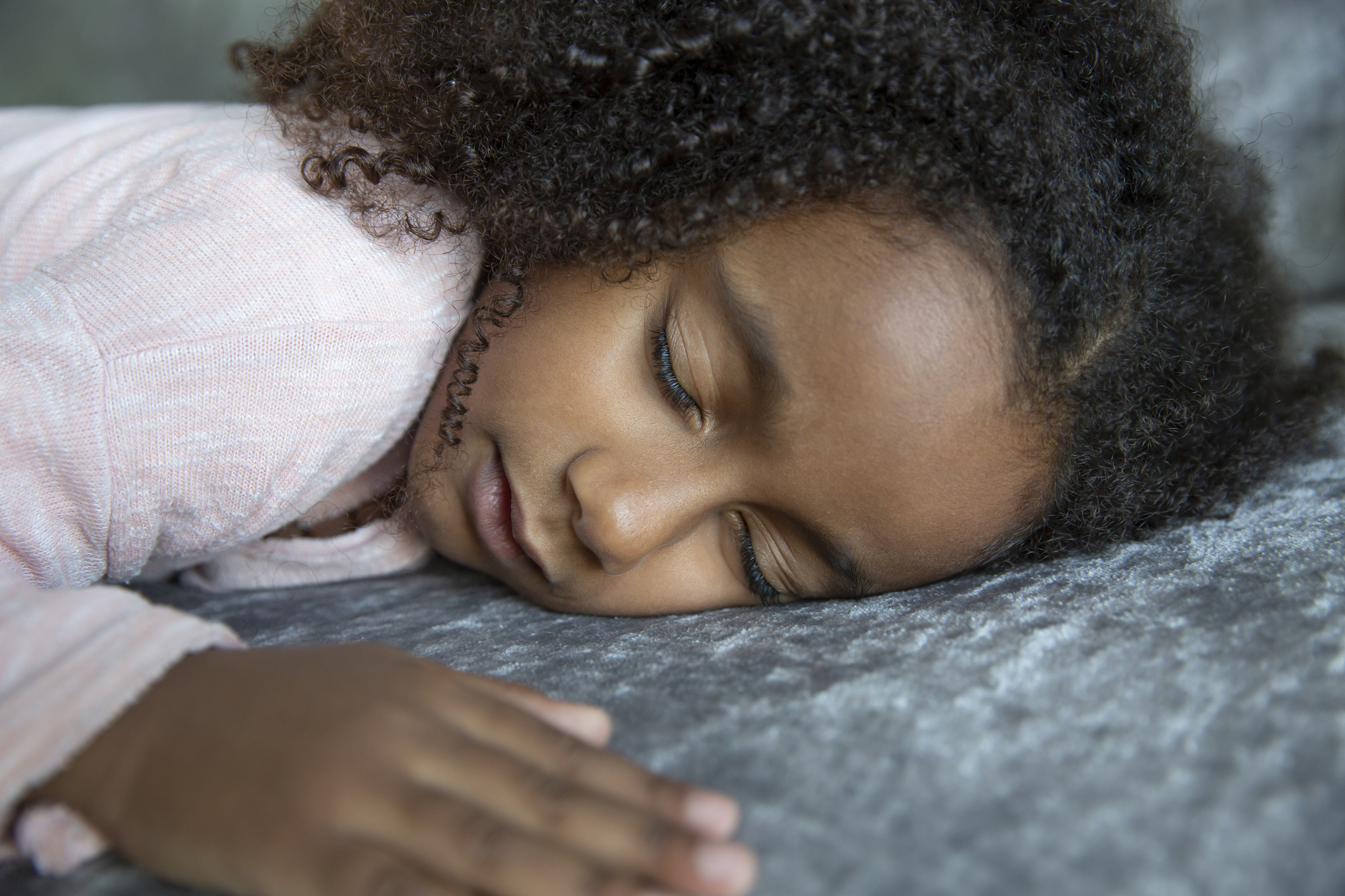 A young child with curly hair is lying down on a soft surface with eyes closed, appearing to be asleep or resting