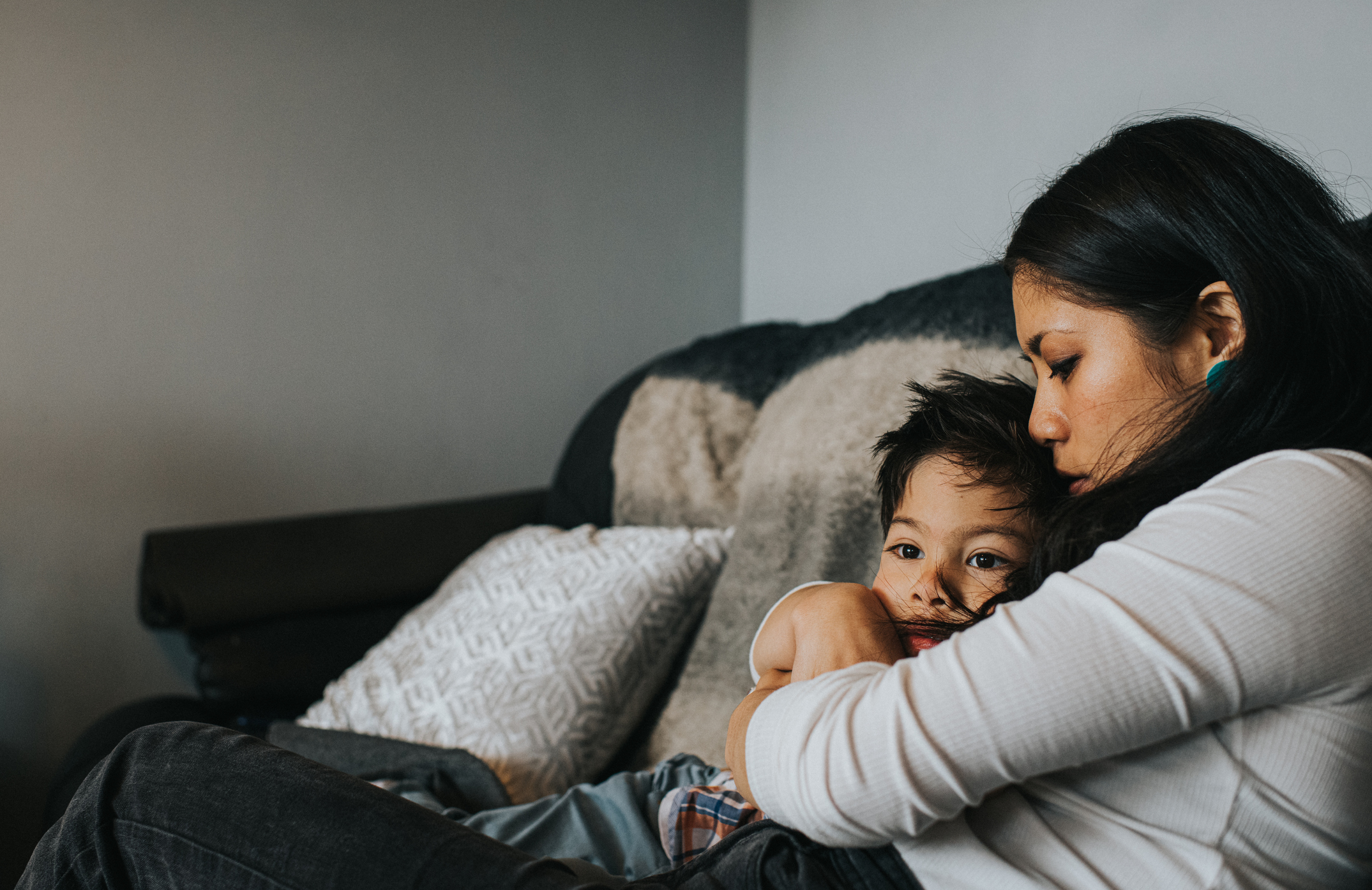 A woman and child sit on a couch, the woman holding the child close as they both look pensive and comfortable