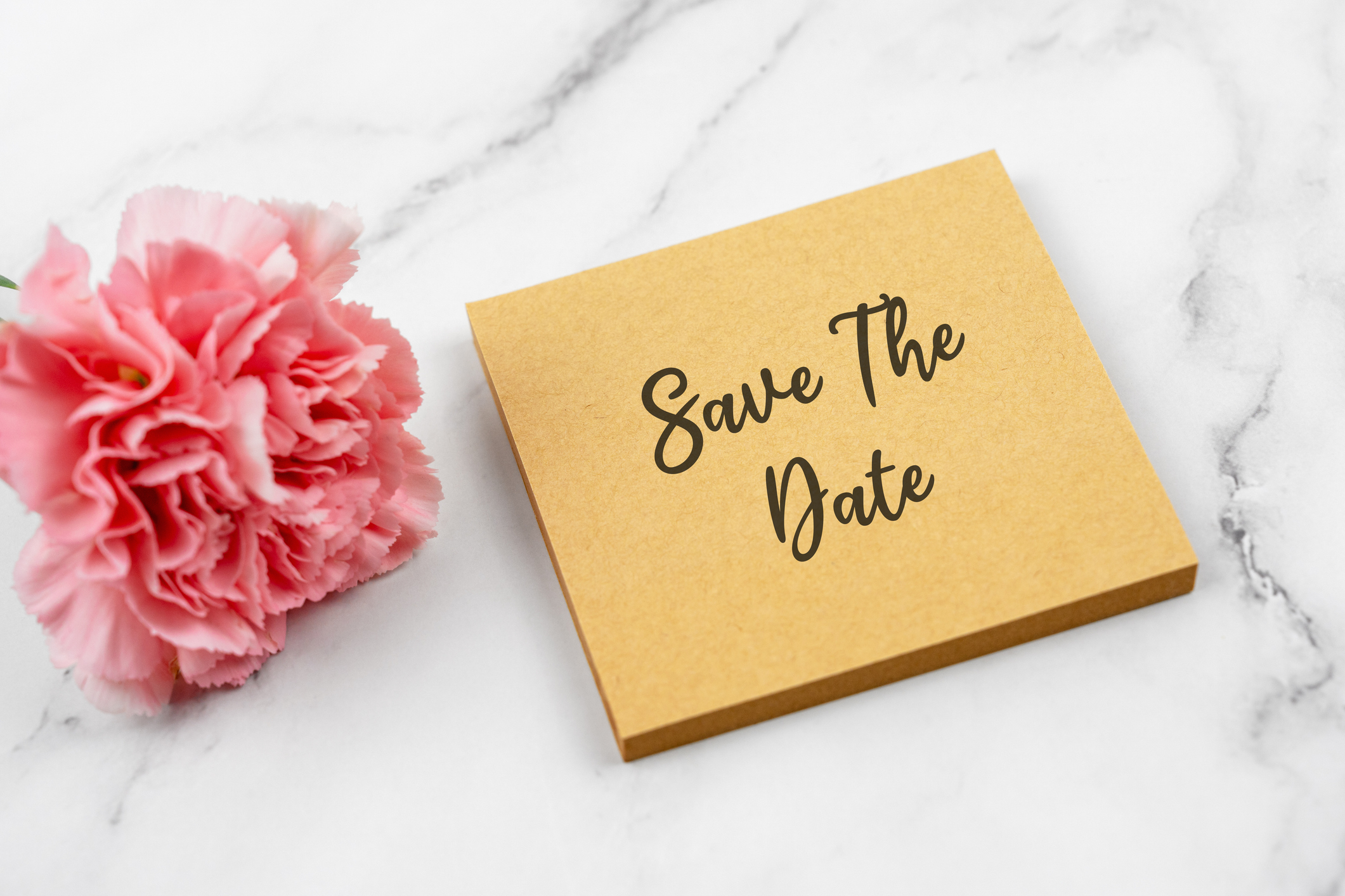 A gold-colored "Save The Date" card is placed on a marble surface next to a pink carnation flower