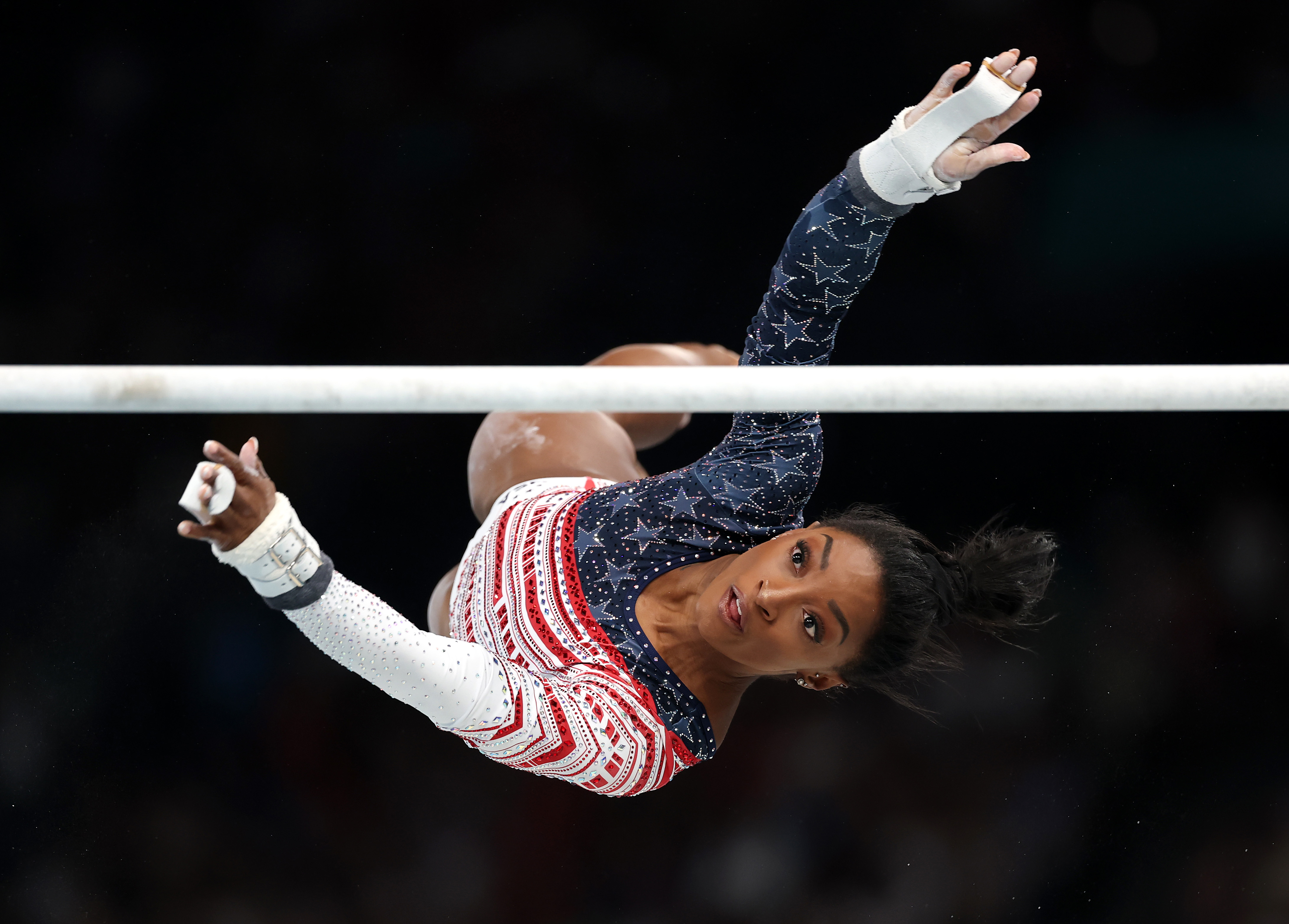 Simone Biles performs a gymnastics routine on the uneven bars, wearing a leotard with star and stripe patterns