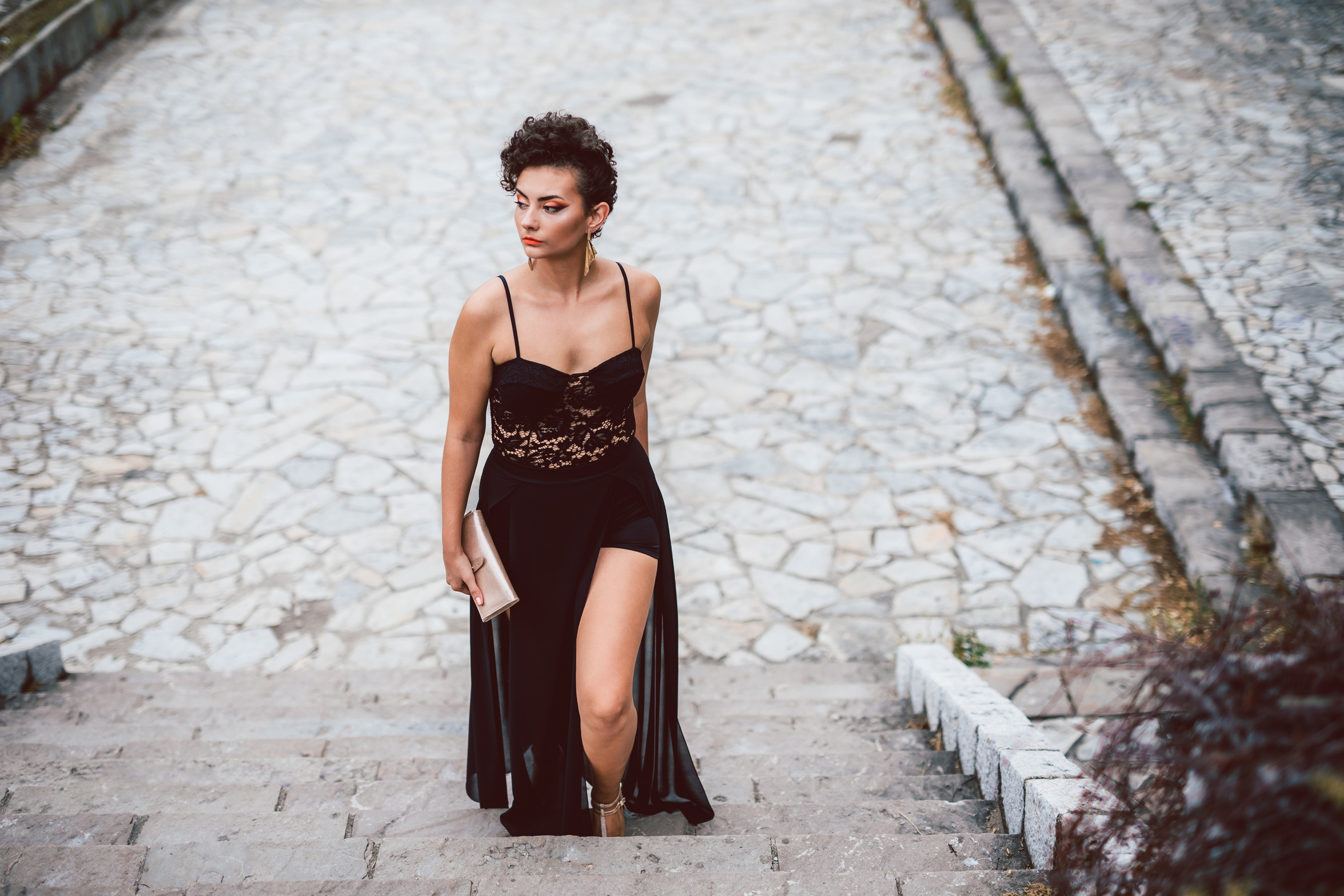 A woman in an elegant black dress with a high slit and lace details walks up stone steps holding a clutch