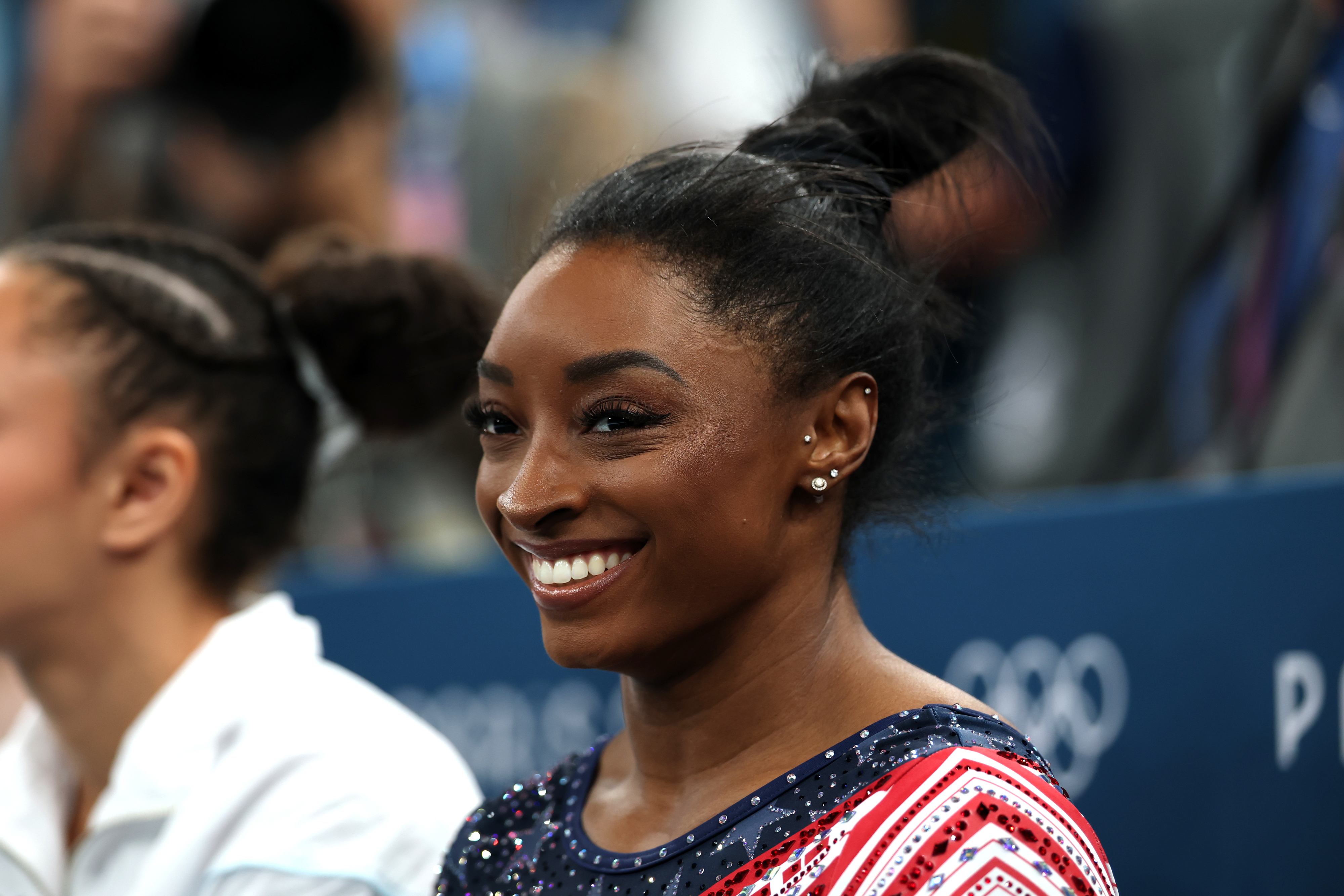 Simone Biles smiles while dressed in a red, white, and blue athletic outfit at what appears to be an Olympic event. A second person is partially visible