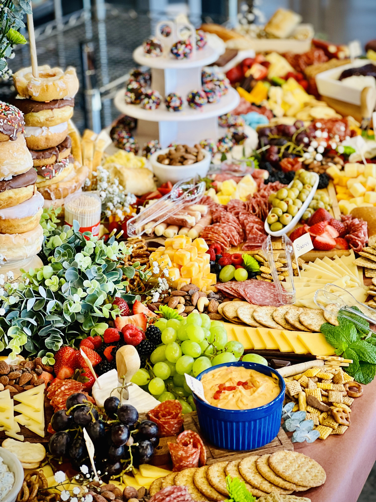 A large, beautifully arranged charcuterie board with a variety of fruits, cheeses, meats, nuts, crackers, and dips, as well as a tiered stand of donuts in the background