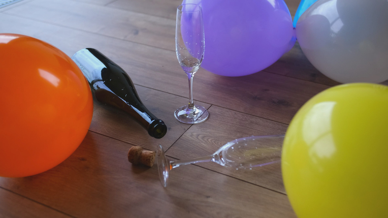 A spilled champagne bottle and glass, surrounded by colorful balloons on a wooden floor