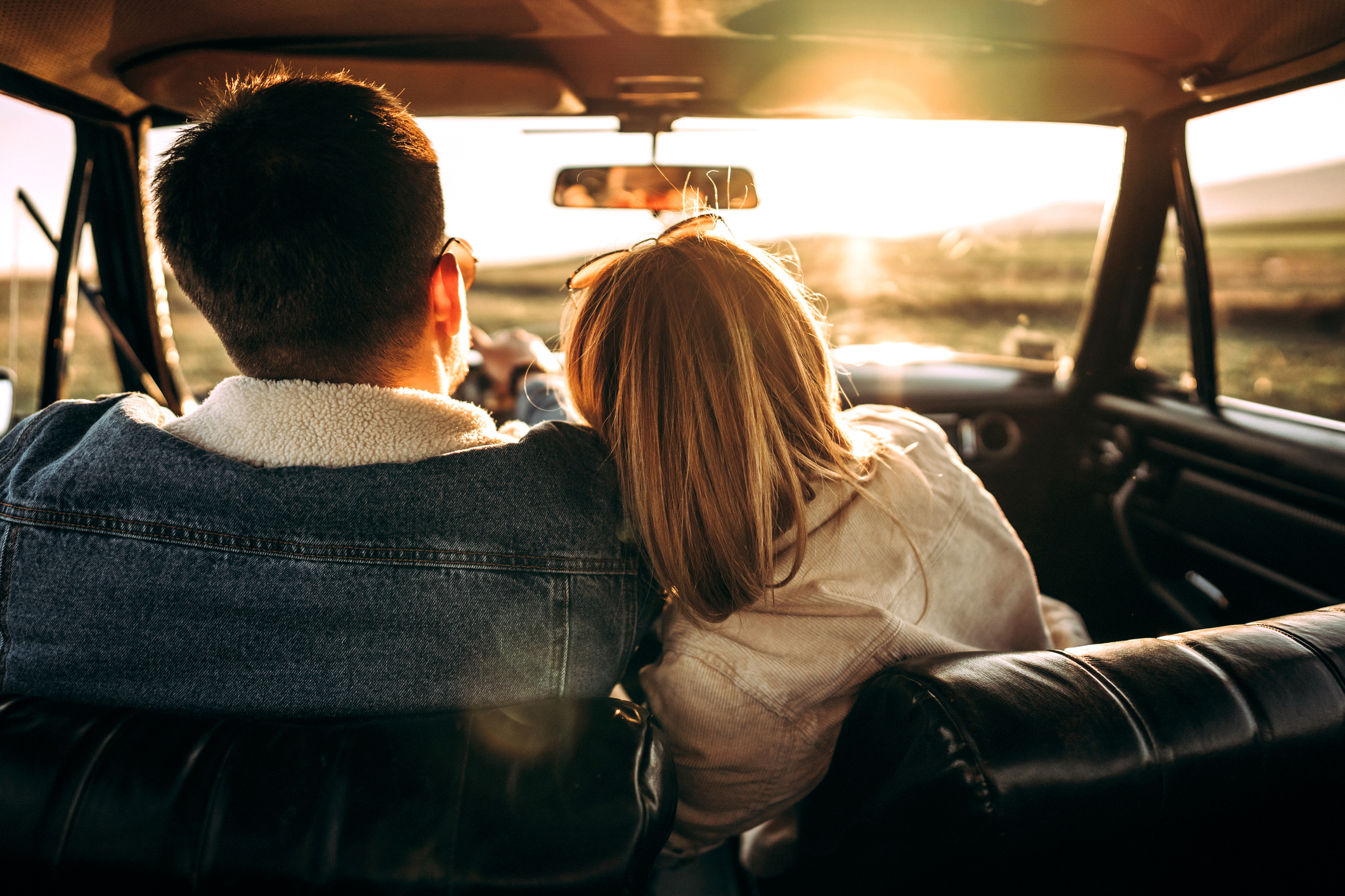 A couple in a car with the woman resting her head on the man's shoulder, looking out at a sunset through the front windshield