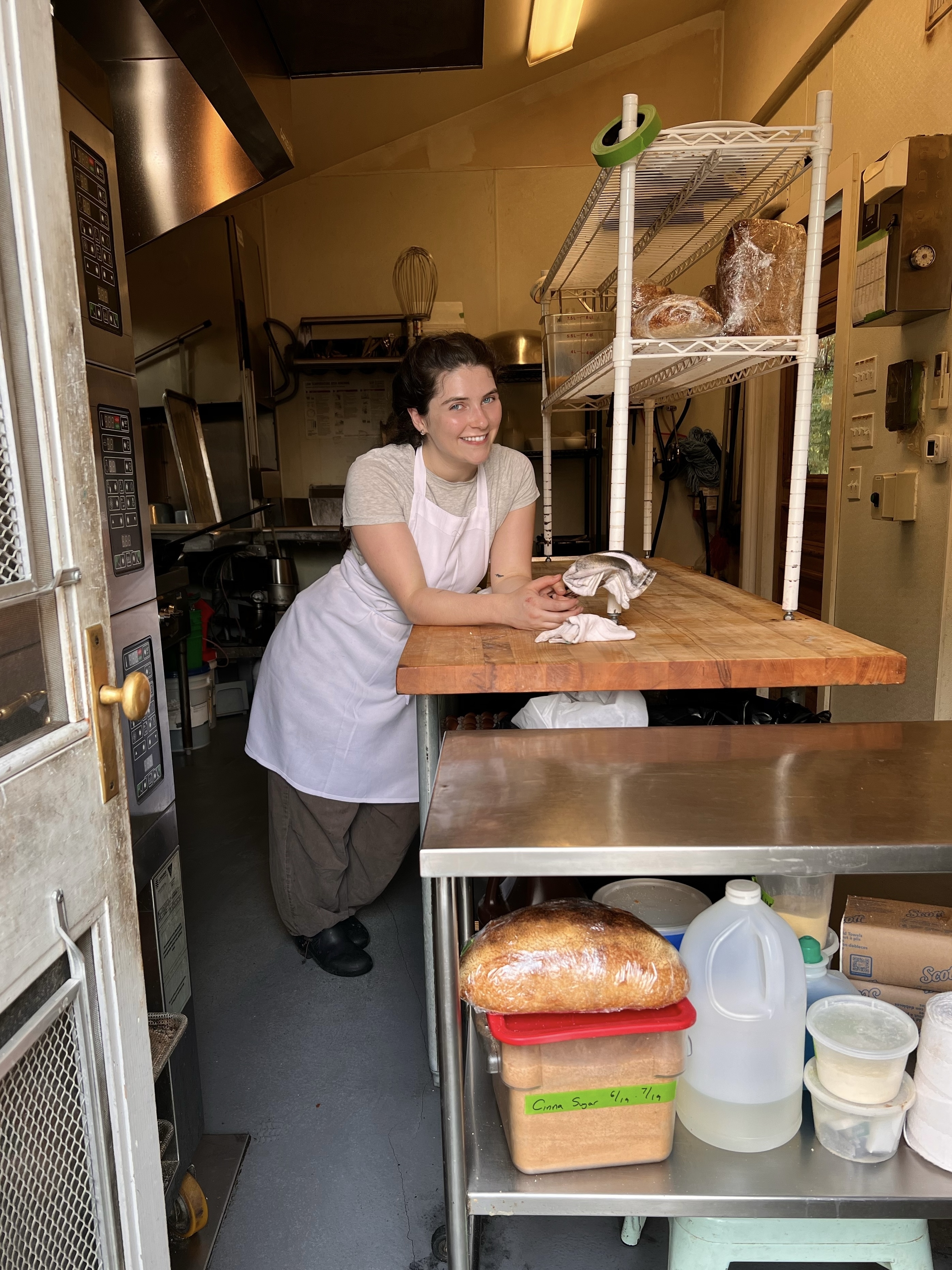 A woman in a kitchen, leaning on a wooden counter with baking ingredients and utensils around her, wearing a white apron and brown pants