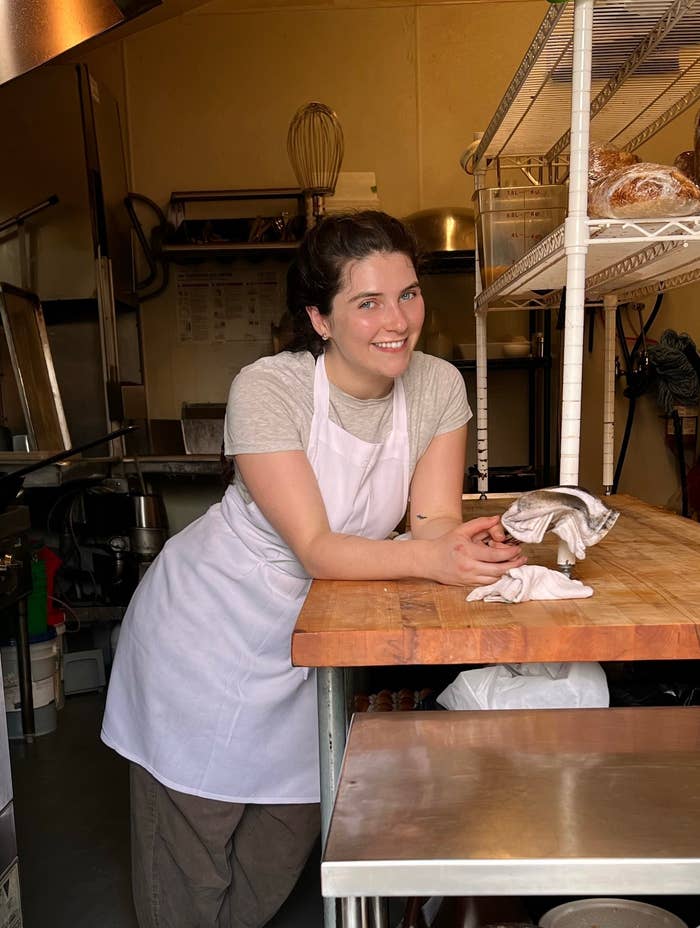 A woman in a kitchen, leaning on a wooden counter with baking ingredients and utensils around her, wearing a white apron and brown pants