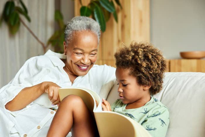 An elder with short gray hair and a child with curly hair share a moment reading a book together on a couch. The elder is smiling while the child is focused on the book