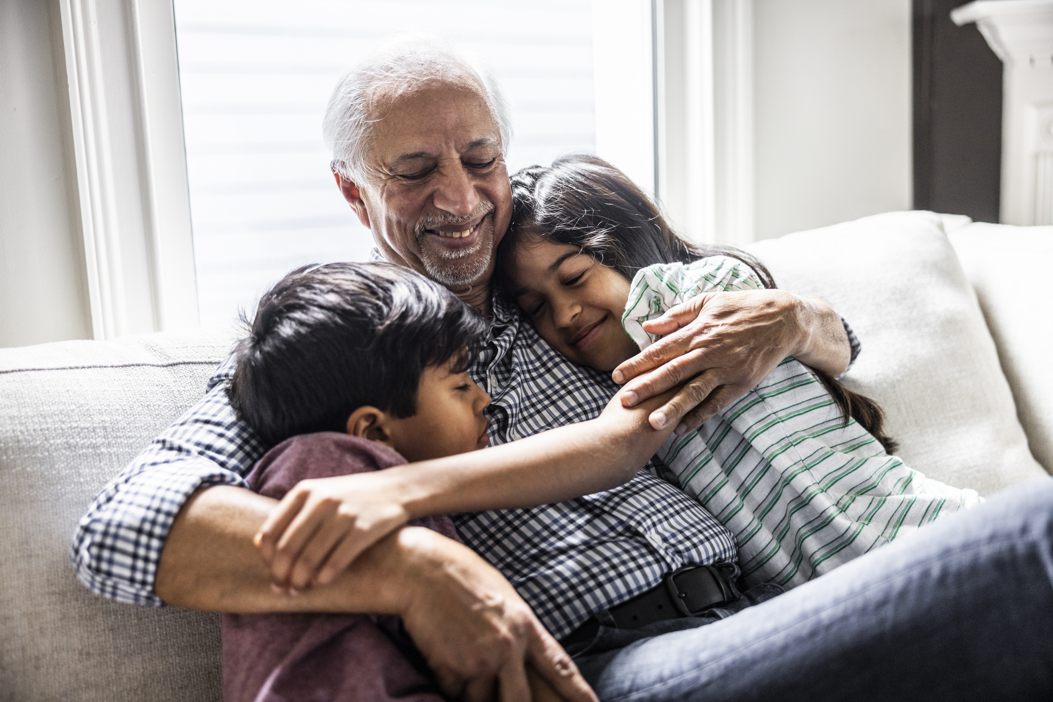 An older man, smiling, sits on a couch hugging two young children who are snuggling close to him