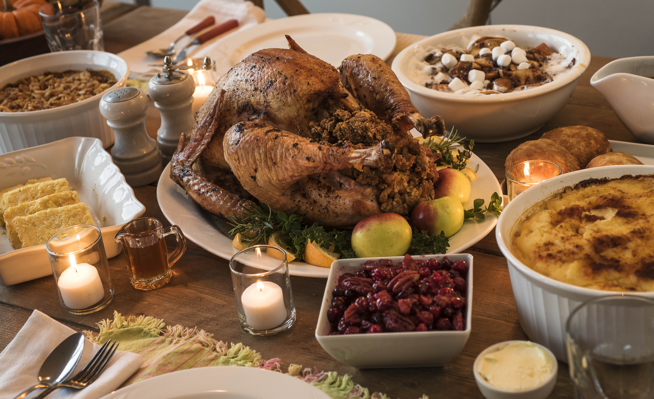 A Thanksgiving dinner table spread with a roasted turkey, stuffing, cranberries, mashed potatoes, cornbread, and various side dishes. Candle jars are lit