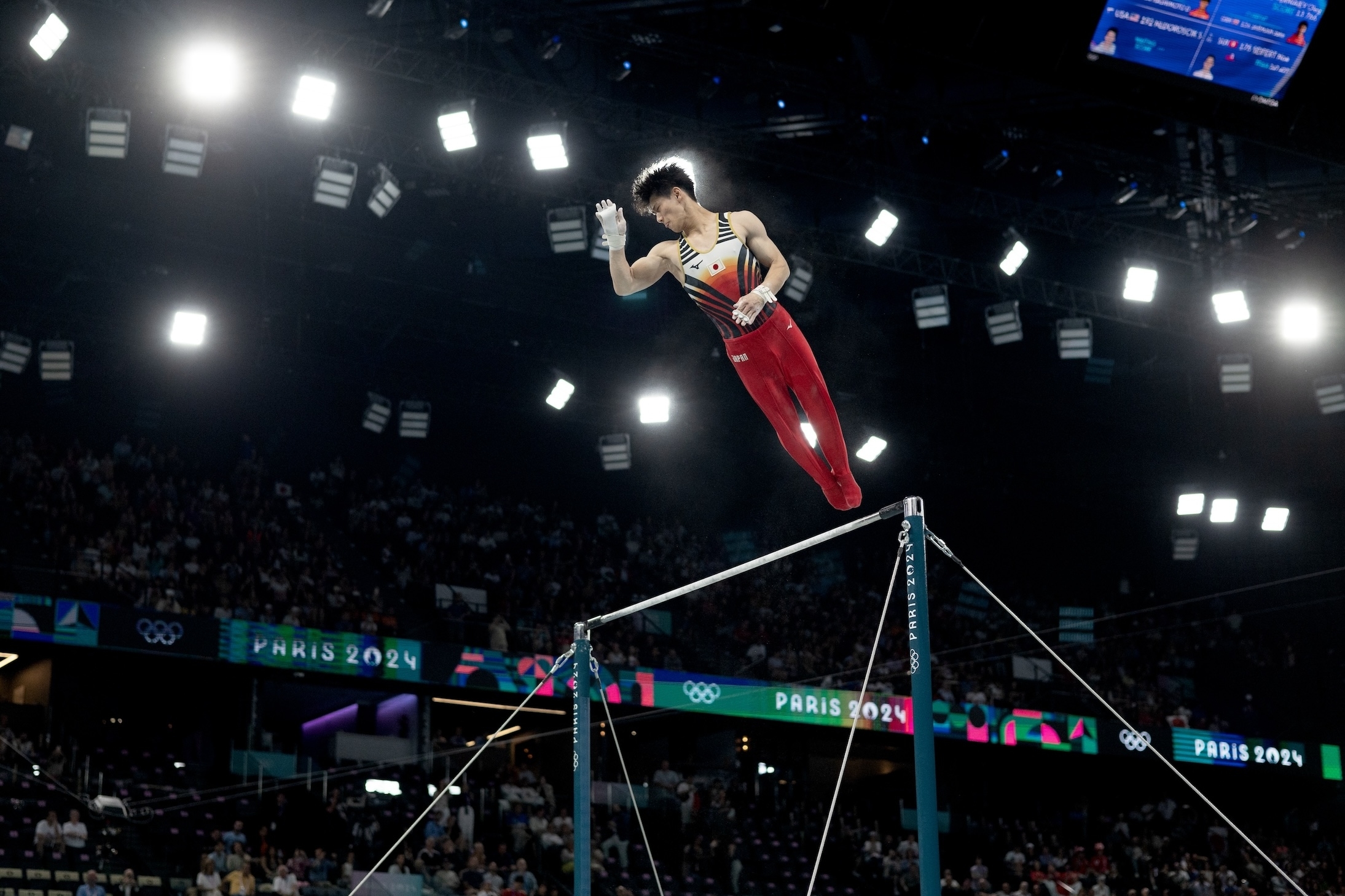 A gymnast performs a routine on the horizontal bar at the Paris 2024 Olympic Games under bright arena lights with an audience in the background