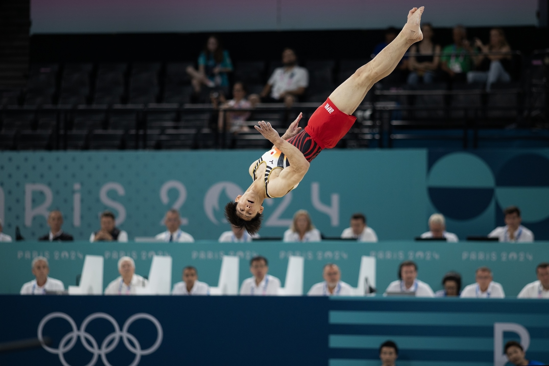 An athlete mid-air during a gymnastics routine at the Olympic Games, judges and spectators watch in the background