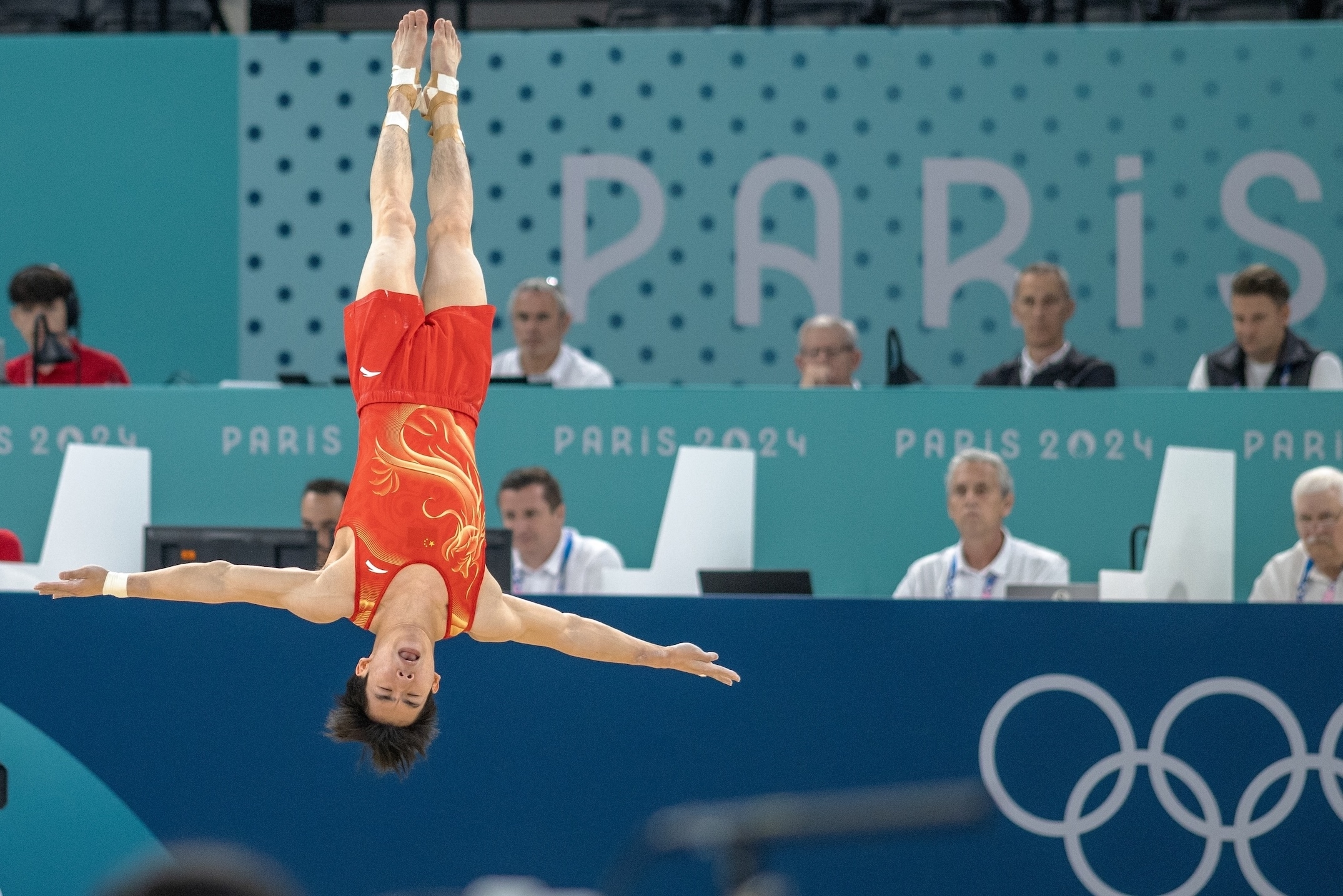 A gymnast in mid-air performing a routine at the Paris 2024 Olympics, wearing a red leotard, with the audience and judges in the background