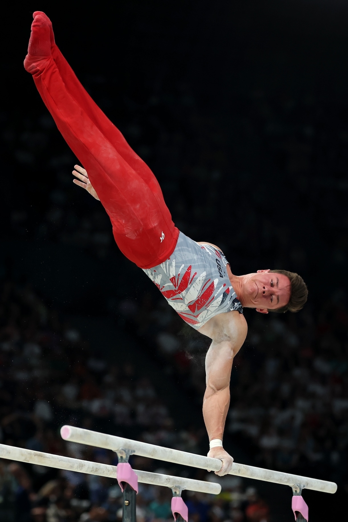 Male gymnast performing a handstand on parallel bars during a competition, wearing a sleeveless top and fitted pants. Name unknown