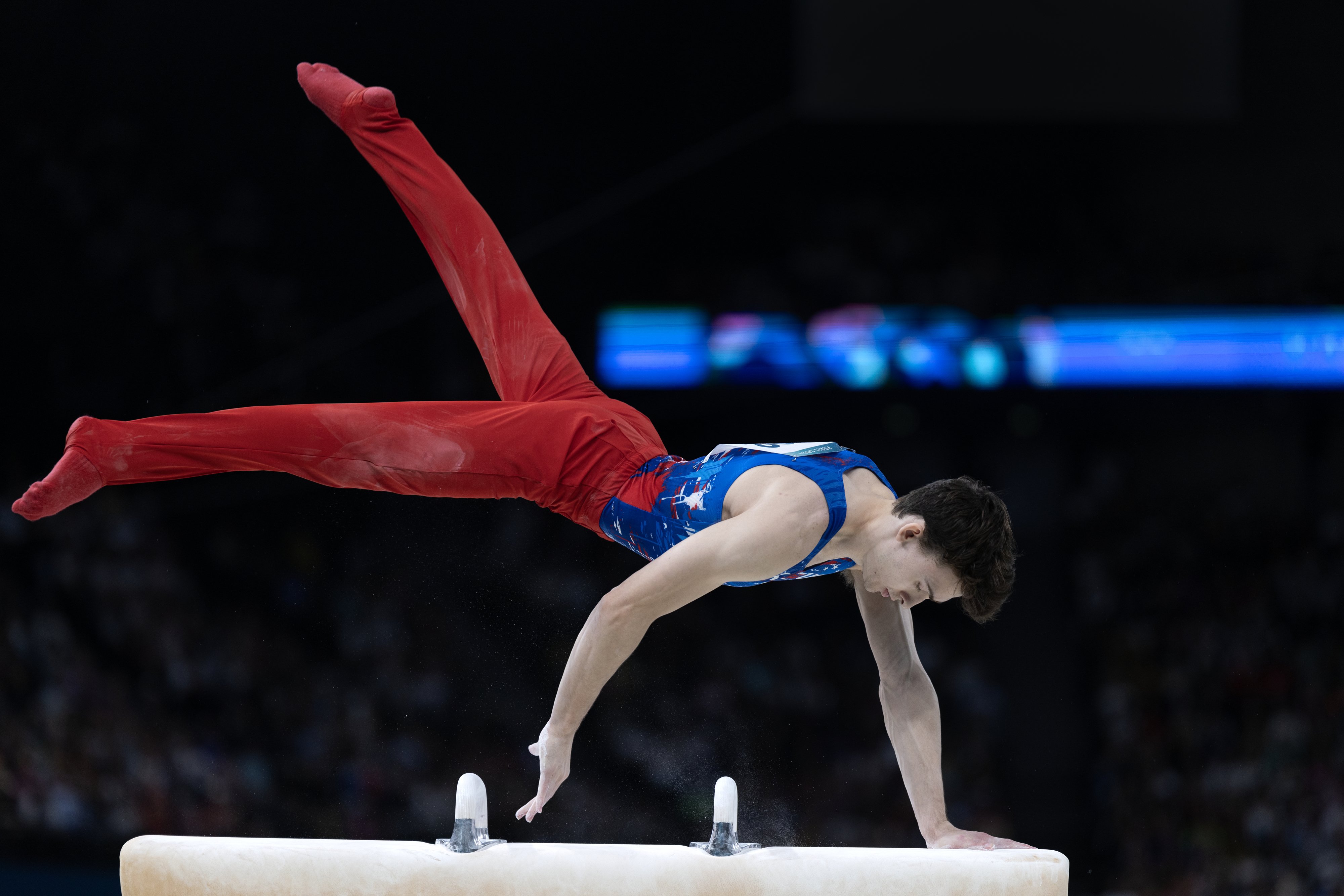 A gymnast performs a pommel horse routine, wearing a blue tank top and red pants, in a crowded stadium