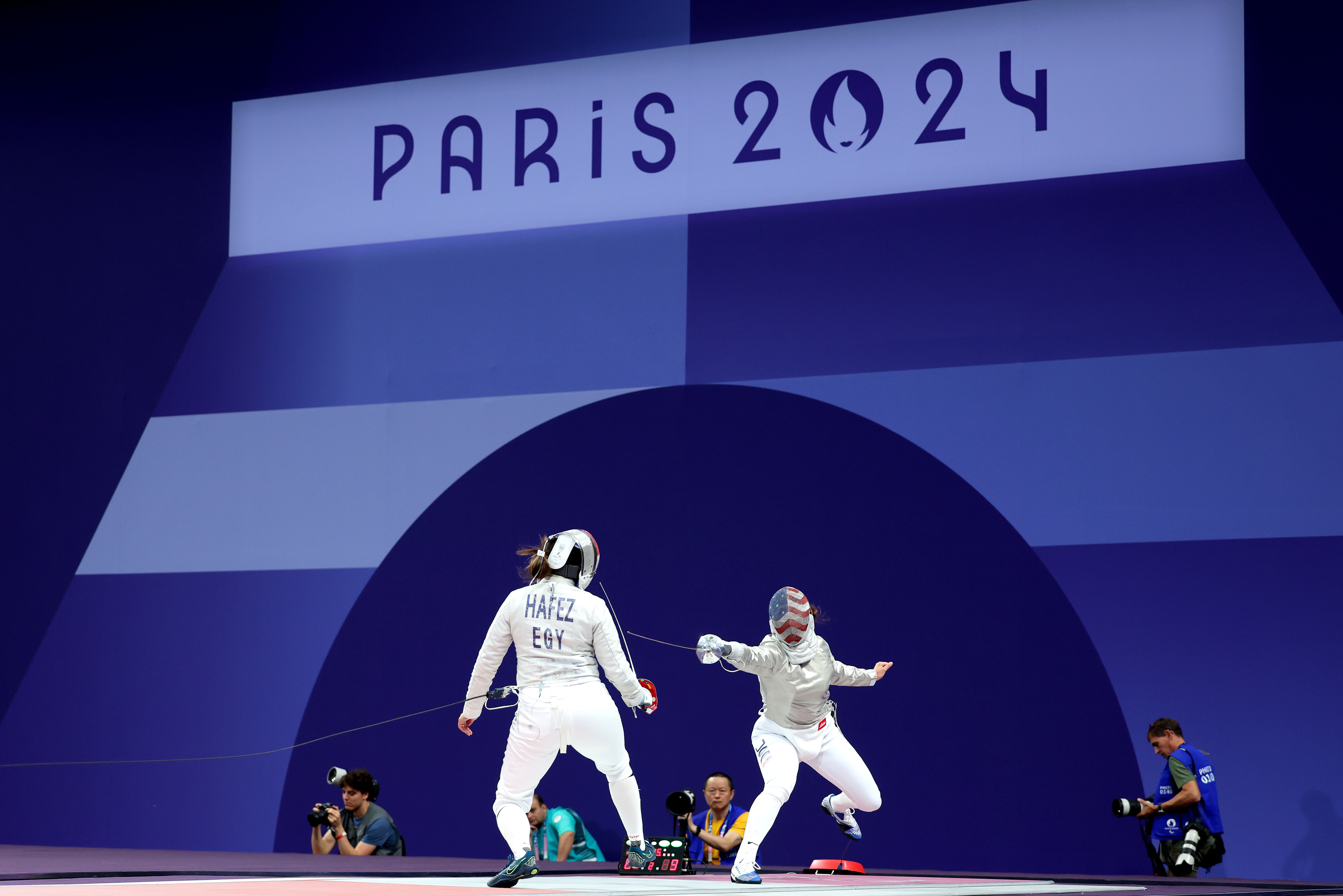 Two fencers compete under a "Paris 2024" sign, while photographers capture the event. One fencer’s suit reads “Hafez EGY.”