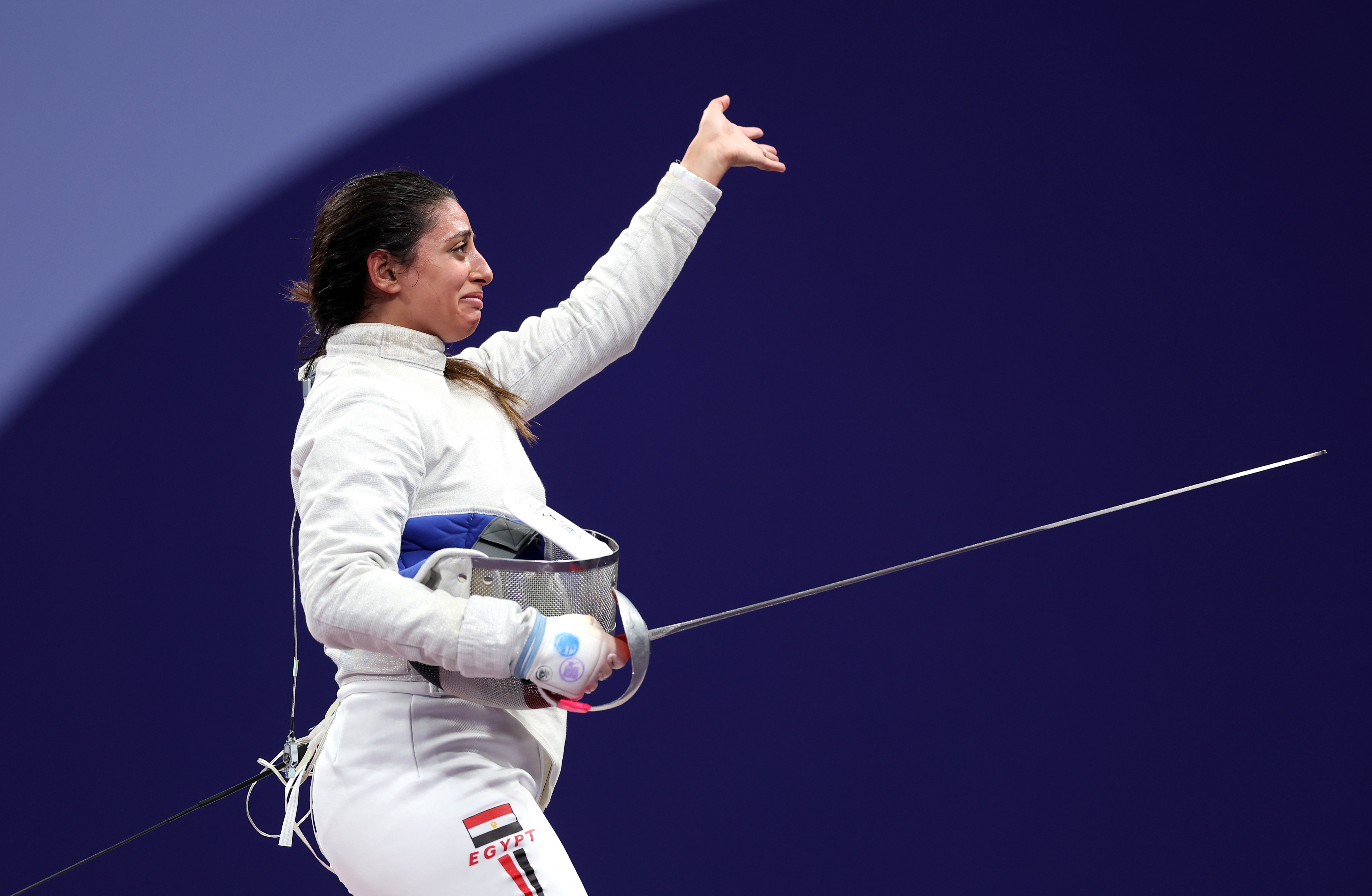 Fencer holding helmet at her hip, raising her arm in greeting. She wears a fencing uniform with "Egypt" written on her pants