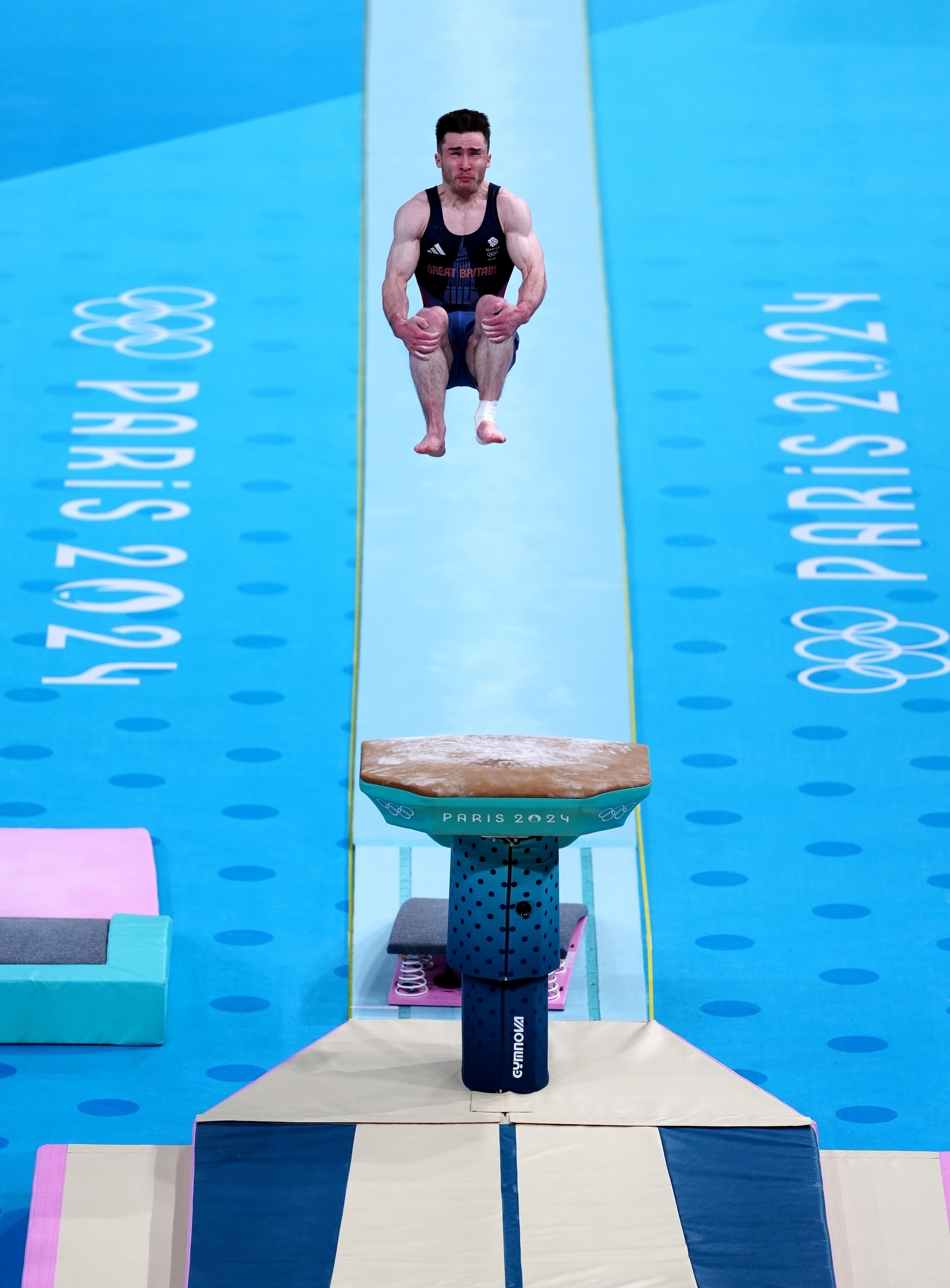 A gymnast is performing a jump on a vaulting horse at the 2024 Paris Olympics. The gymnast is mid-air with knees bent, in a focused posture