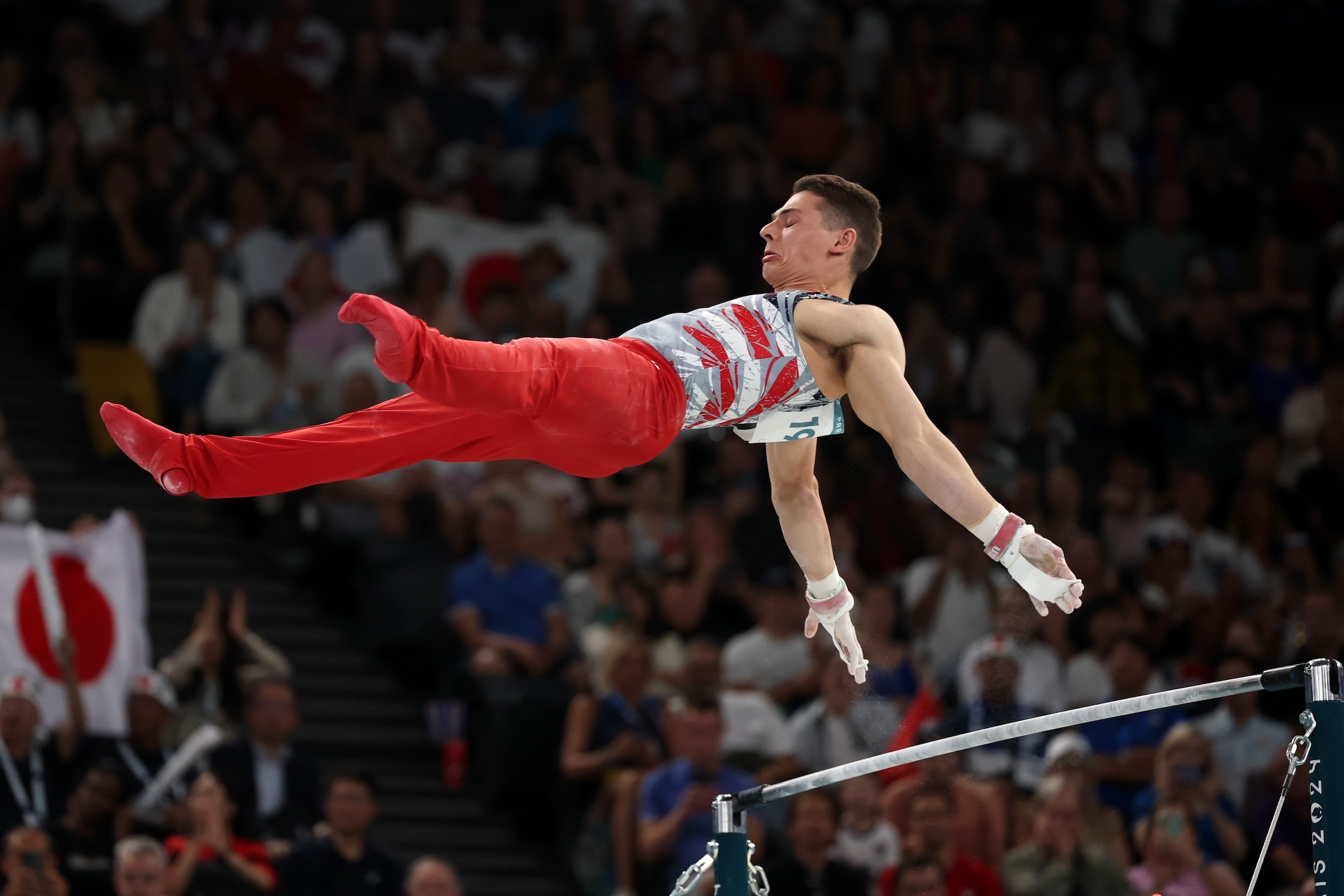 Gymnast Artur Dalaloyan performs a high bar routine during a crowded competition, appearing airborne in a dynamic pose