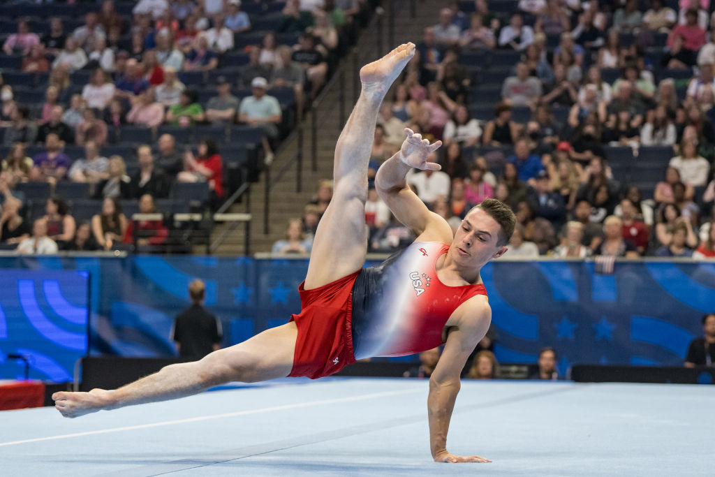 The image shows gymnast performing a floor routine in front of a large audience in an indoor arena, wearing a red and white uniform