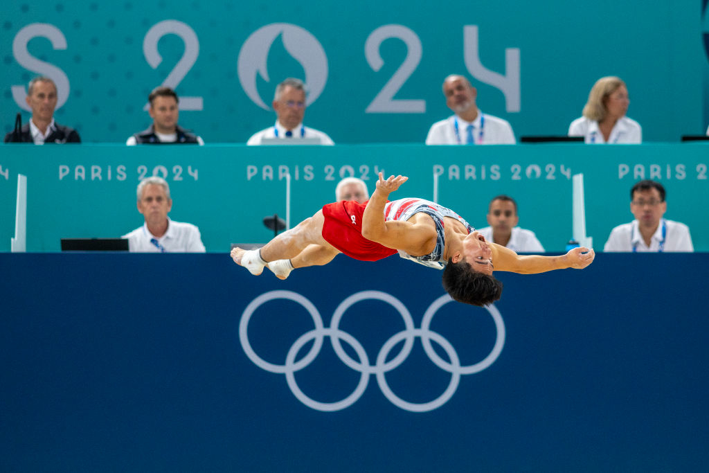 A gymnast in mid-air performs a flip during the Paris 2024 Olympics in front of judges. Olympic rings are visible in the background