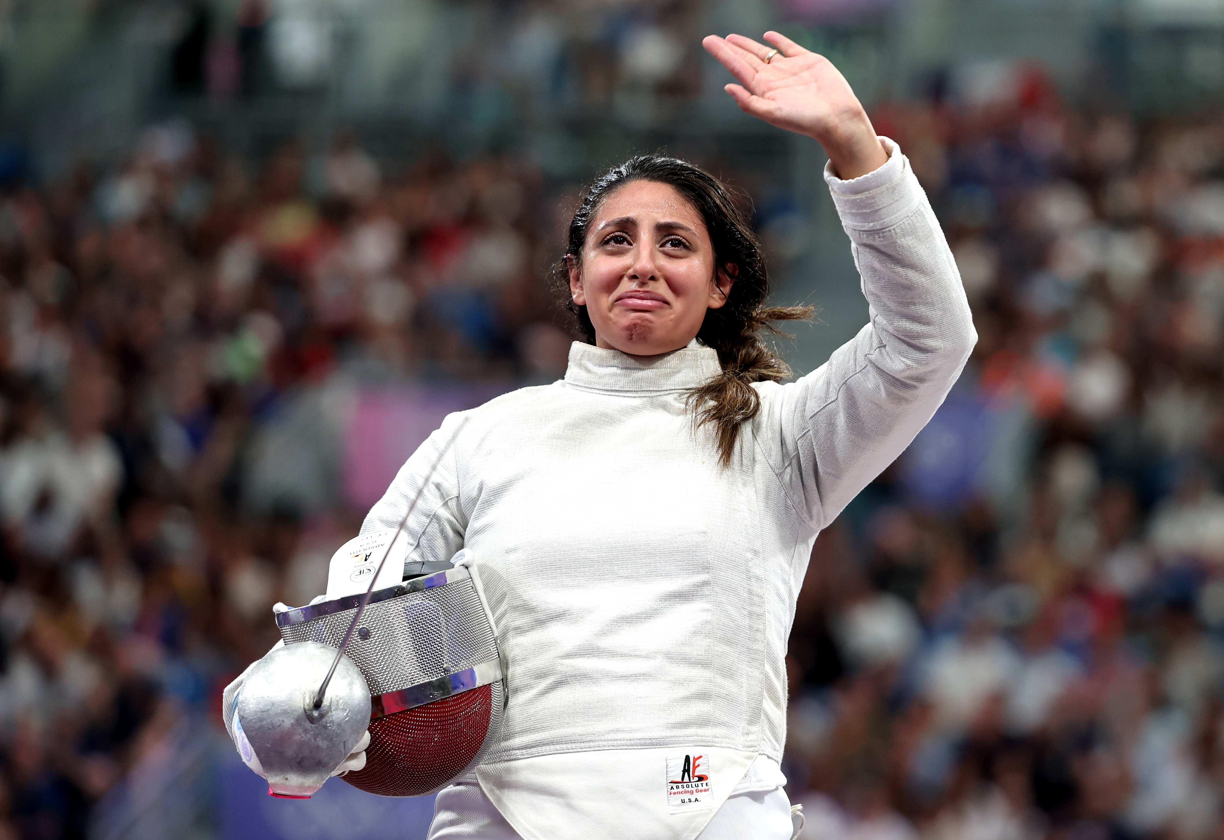 A fencer in full gear, holding her mask, waves to the crowd with a tearful expression