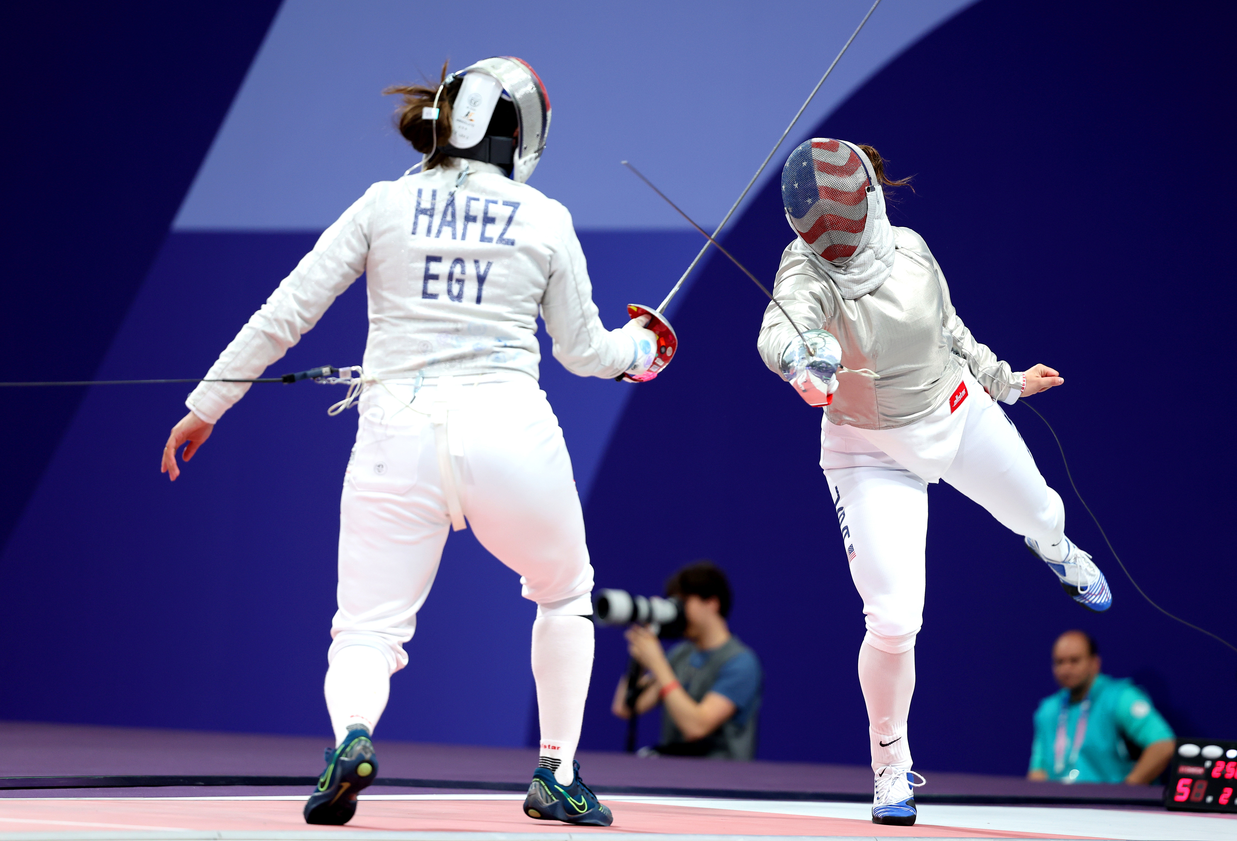 Two fencers, one named Hafez from Egypt, in full fencing gear competing in a match. A photographer and two other individuals in the background