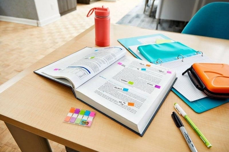 Open textbook with colorful sticky notes, highlighters, pens, a water bottle, a notebook, and a folder on a wooden table, ideal for back-to-school shopping