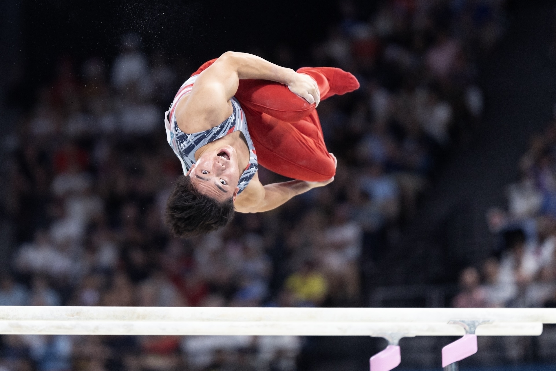 Gymnast performing a high-flying skill on the parallel bars at a competition, wearing a patterned tank top and red pants. Audience in the background