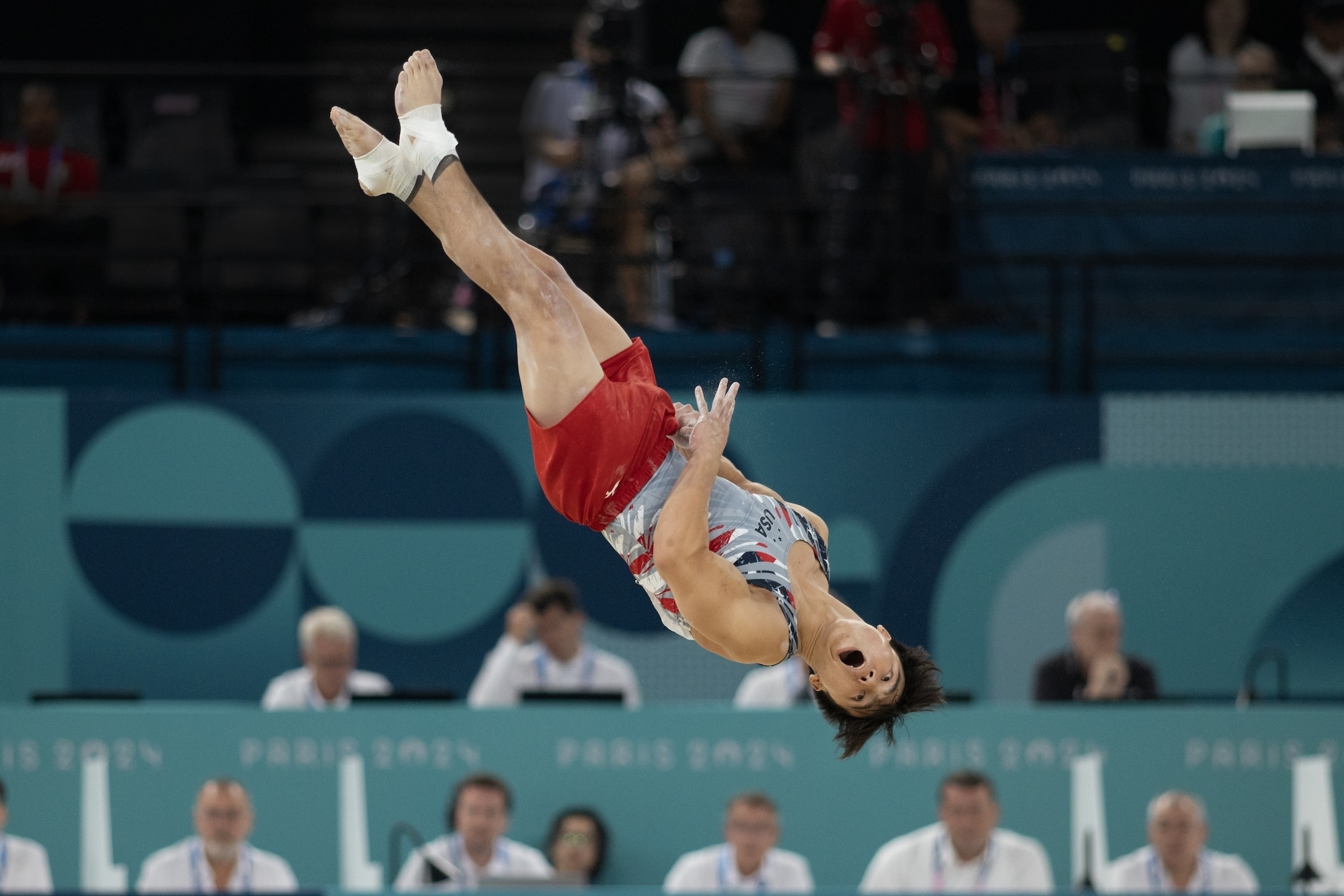A gymnast performs a high-flying flip during a competition, with judges observing in the background