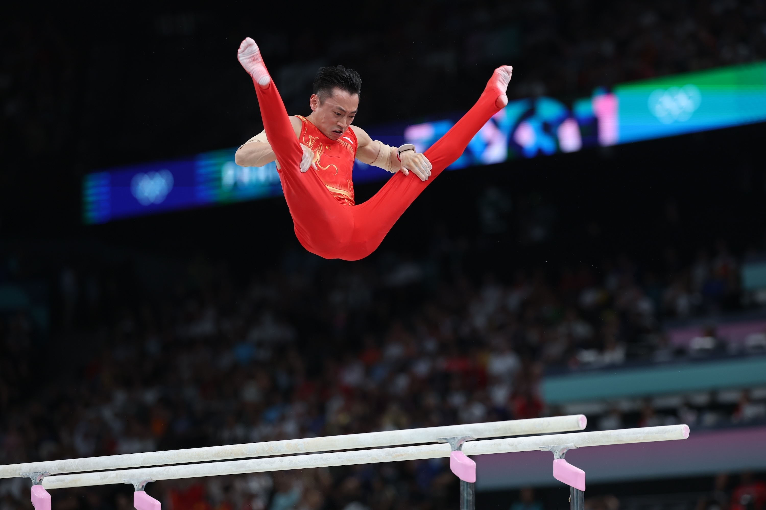 Gymnast dressed in red performs a split jump on parallel bars during a competition, surrounded by a large audience