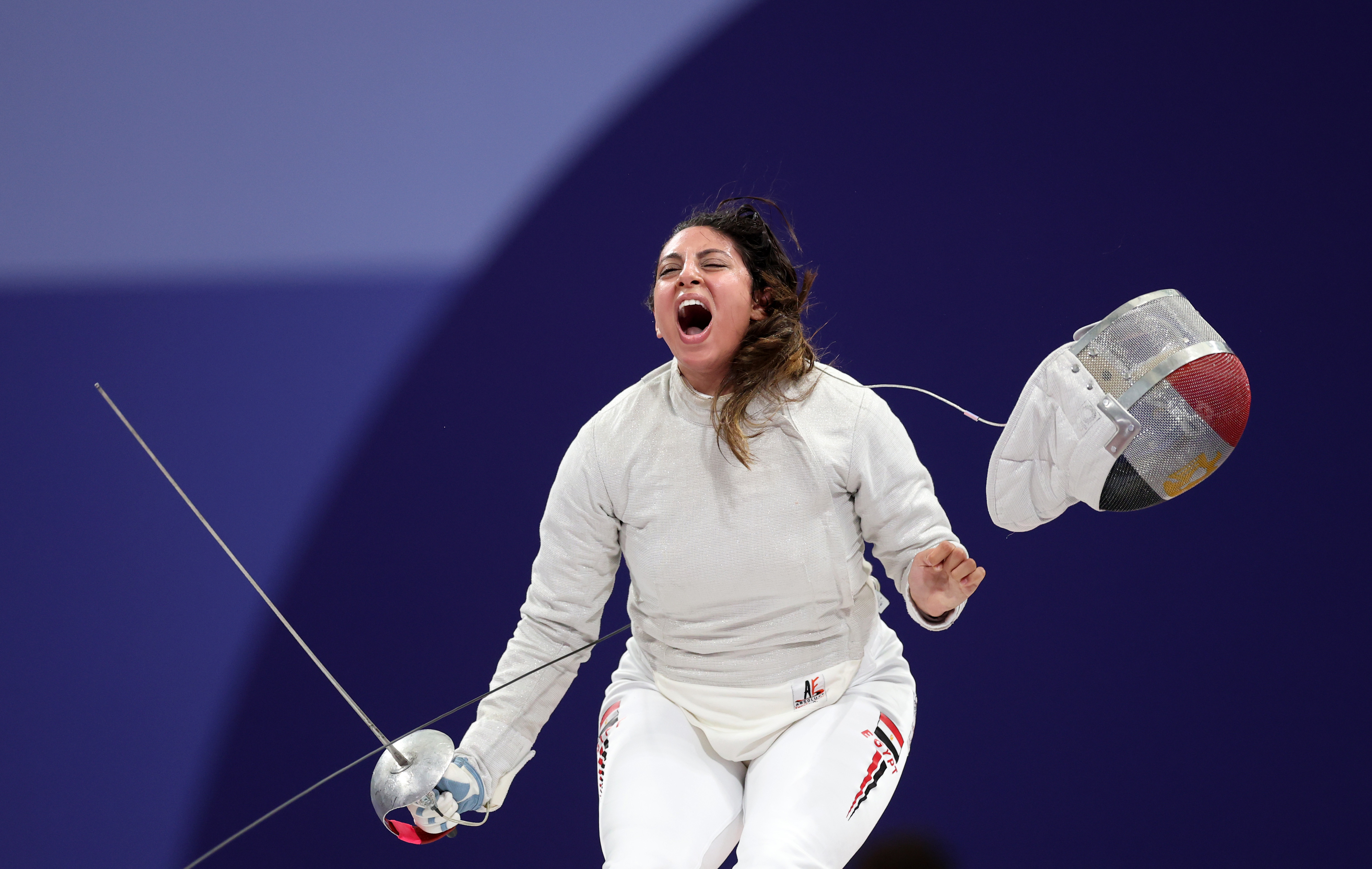 A fencer, wearing full fencing gear and holding a saber, celebrates with an open mouth after a match victory
