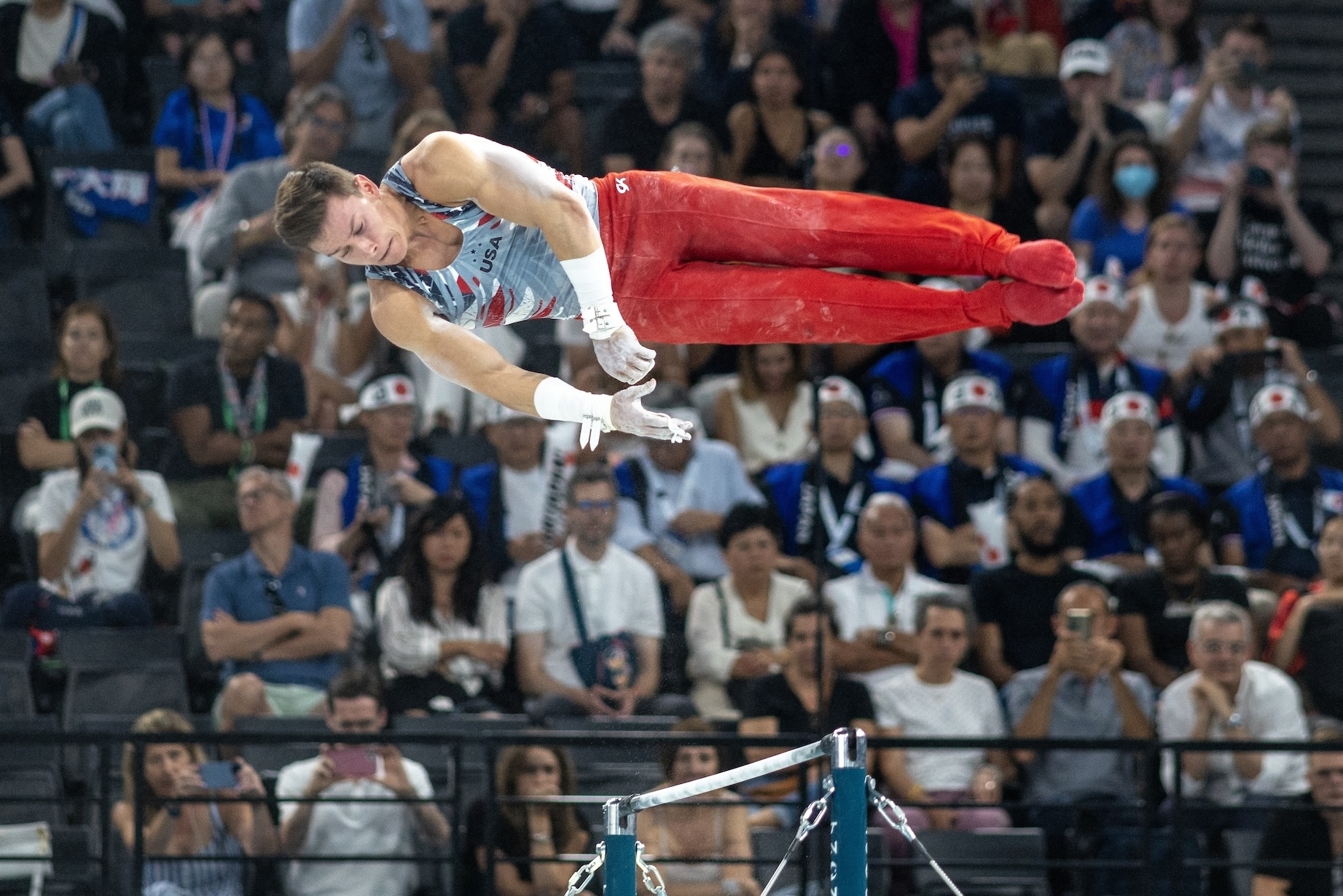 A gymnast performs a horizontal bar routine at a competition with spectators watching in the background