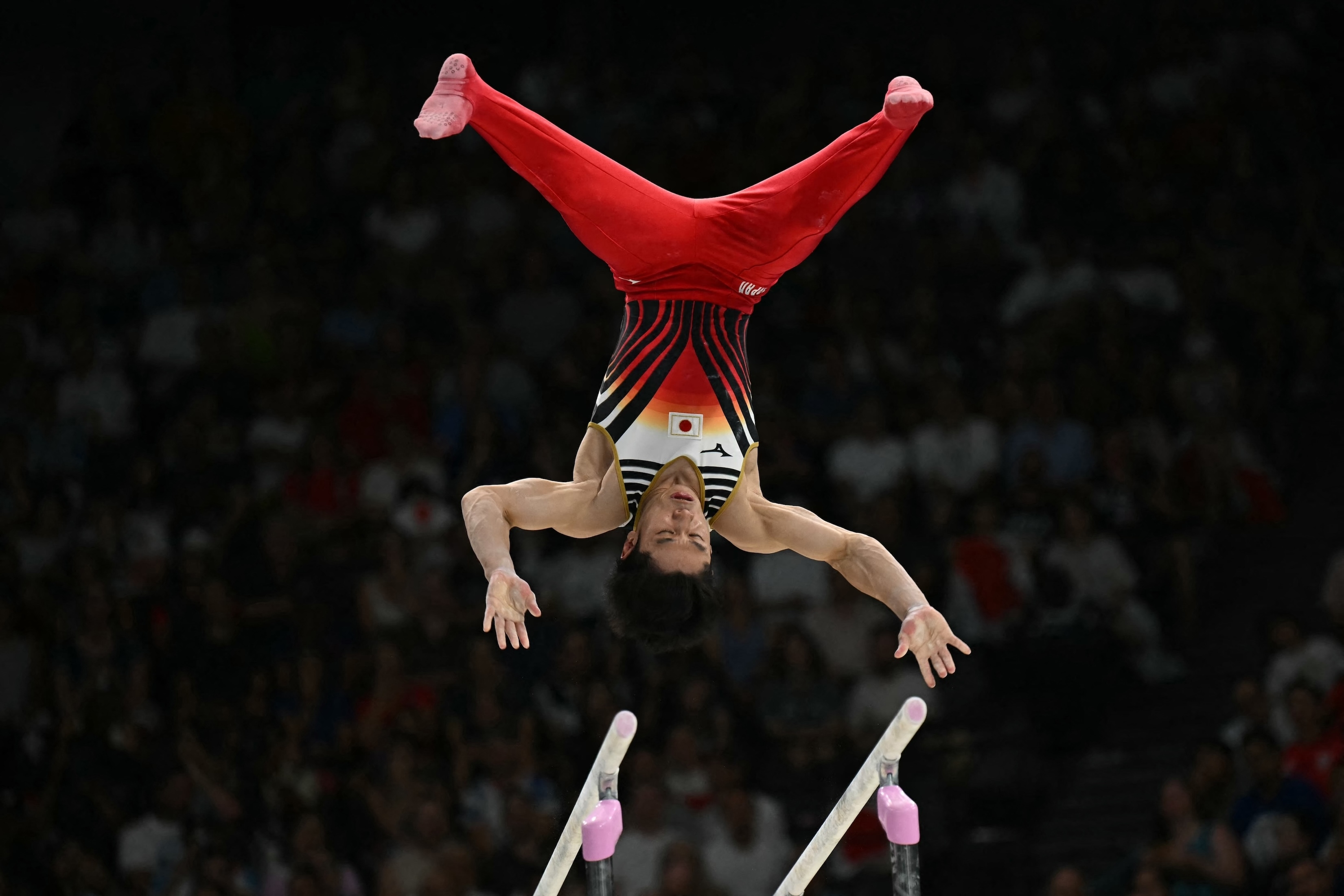 Japanese gymnast performing an upside-down parallel bars routine, wearing a leotard with red, white, and black stripes, in front of a full audience