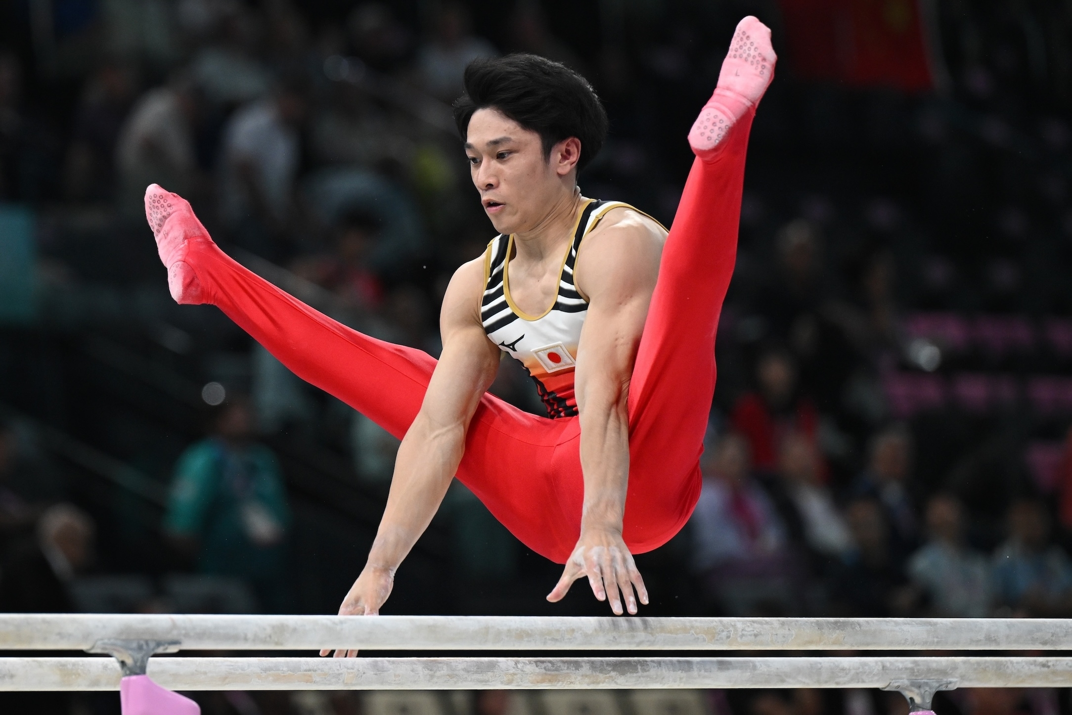 Gymnast performing a routine on the parallel bars, wearing a sleeveless top and red pants. The person is in an airborne straddle position